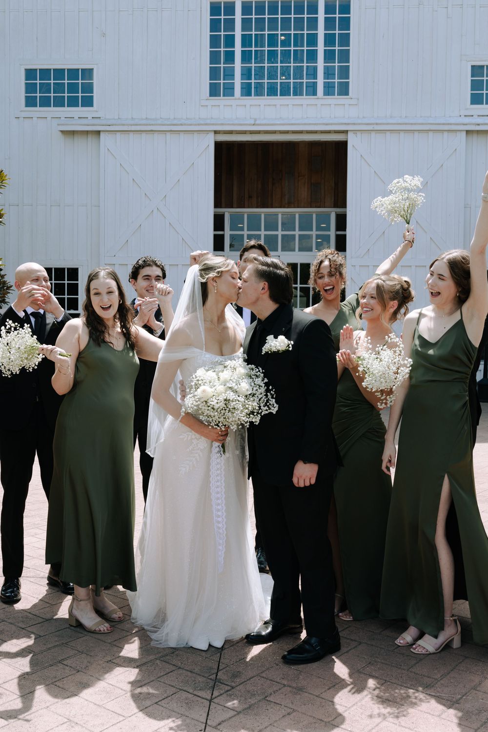 Wedding party celebrating outside a white barn with bridesmaids in dark green dresses holding white bouquets.