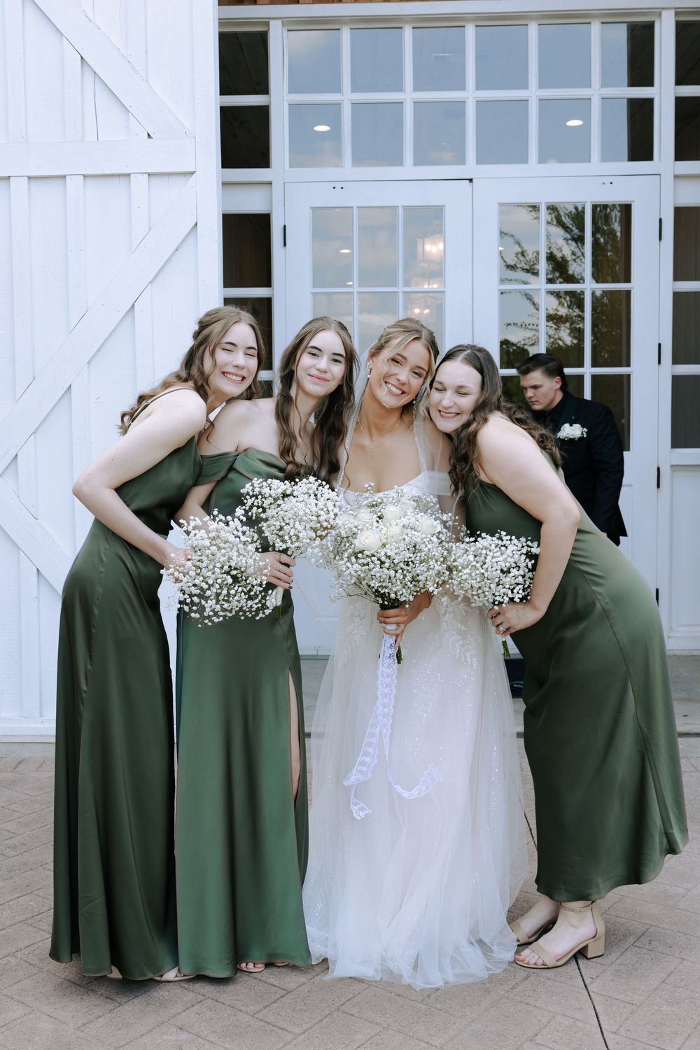 Bridesmaids in elegant sage green dresses pose with bride holding baby's breath bouquets.