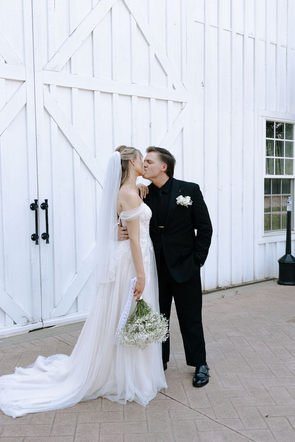 A couple shares a romantic moment in front of a white barn door, with the bride holding a bouquet of white flowers.