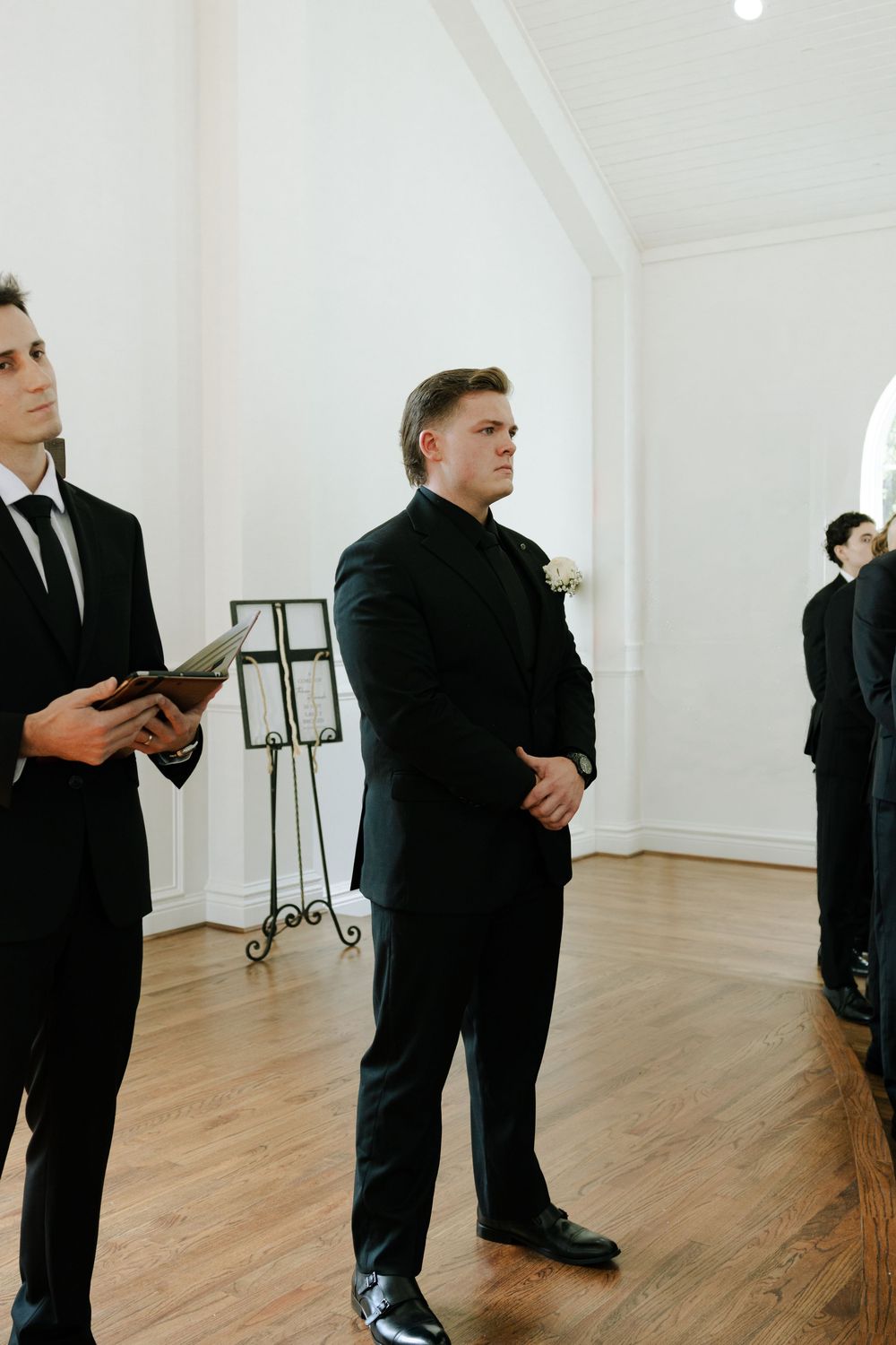Three suited attendants stand in a minimalist white room with hardwood floors during a formal event.