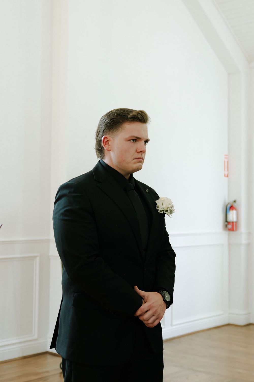 Person in formal black suit with boutonniere awaits in white chapel interior.