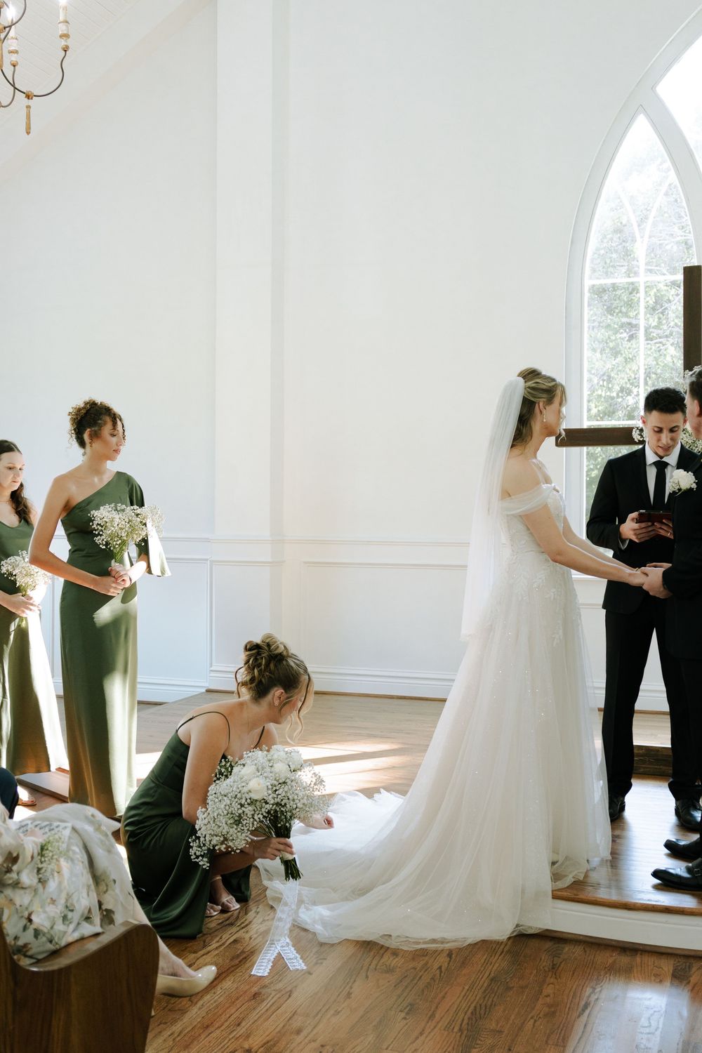 Wedding ceremony taking place in a bright white chapel with bridesmaids in sage green dresses and classic architectural details.