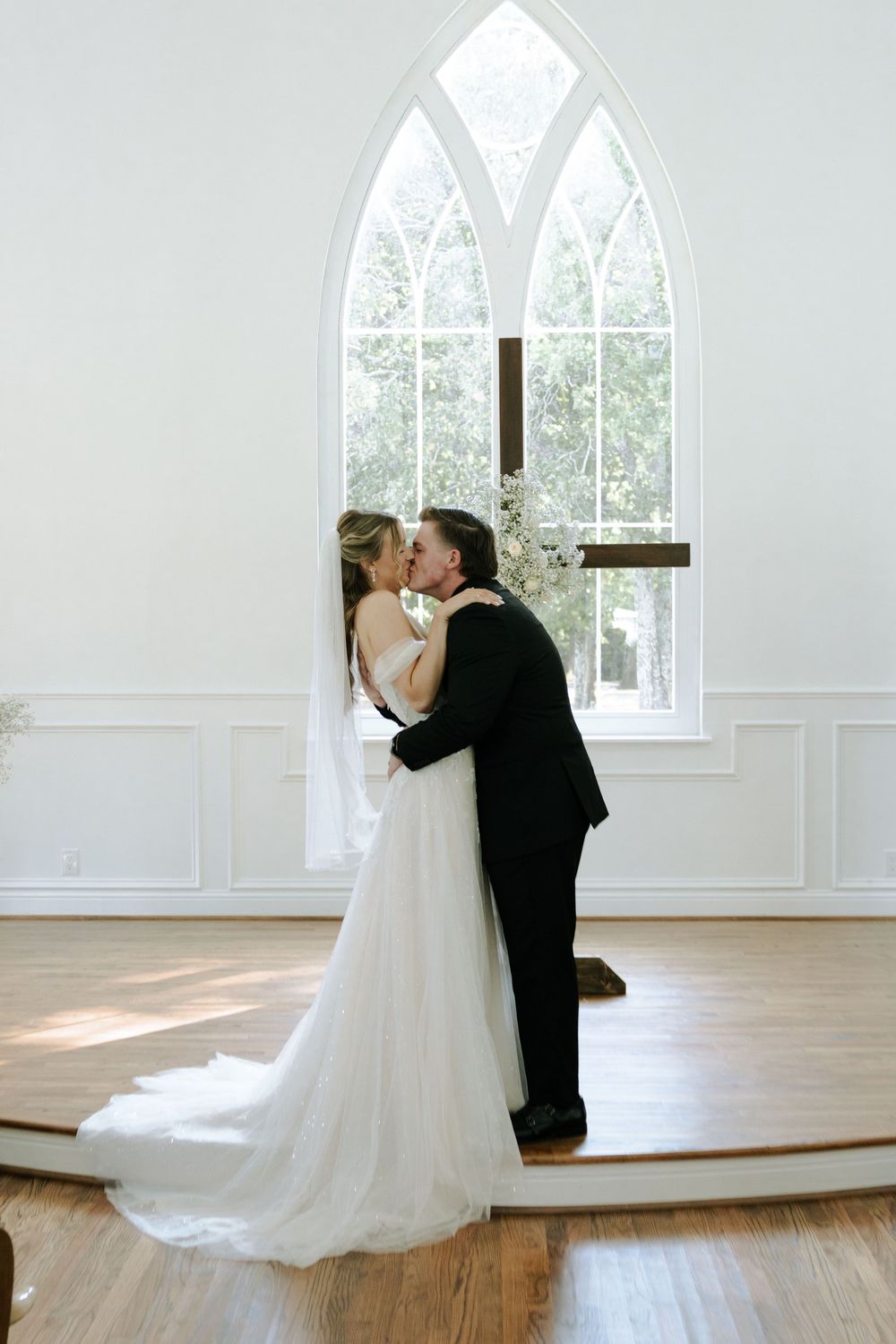Intimate wedding moment captured in front of a gothic arched window in a white chapel.