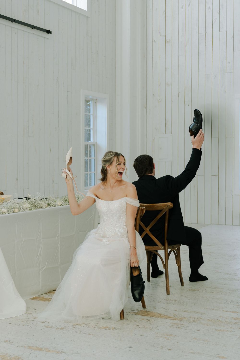Newlyweds playing a wedding reception game while sitting in wooden chairs in an all-white venue.