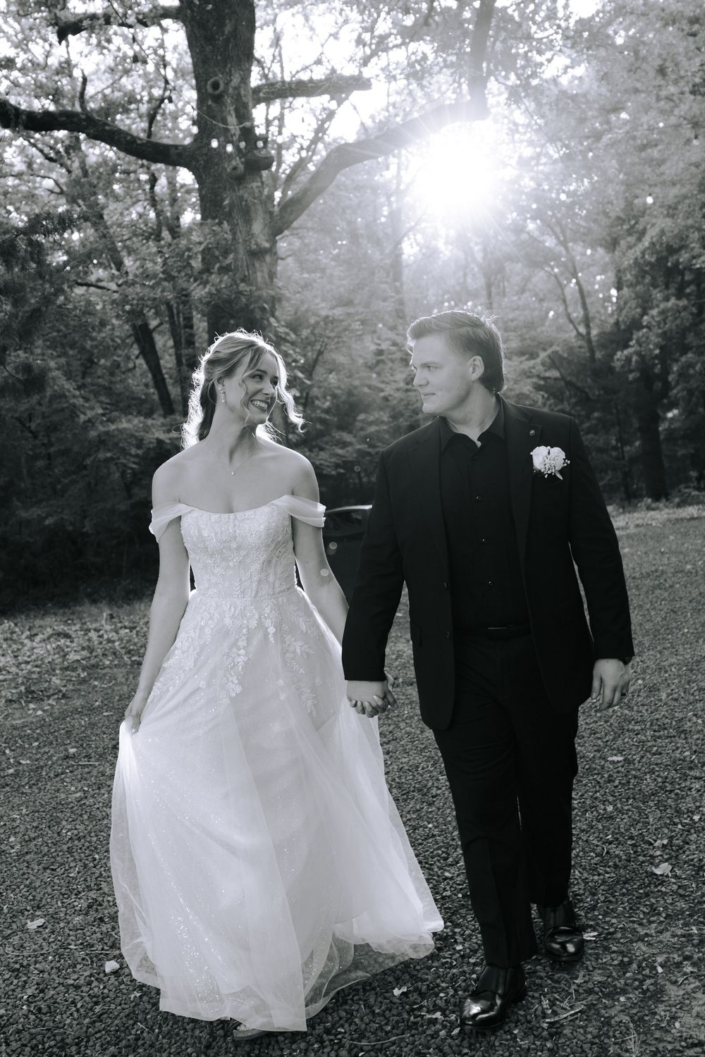 A couple in wedding attire walks through a misty wooded area, with sunlight filtering through the trees in black and white.