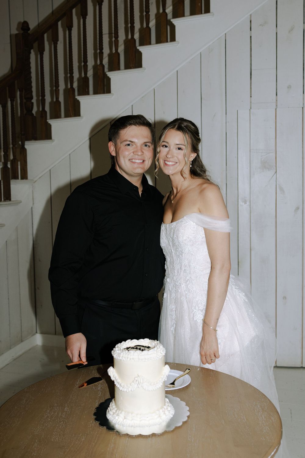 A couple stands beside their white wedding cake in front of a wooden staircase at an indoor celebration.