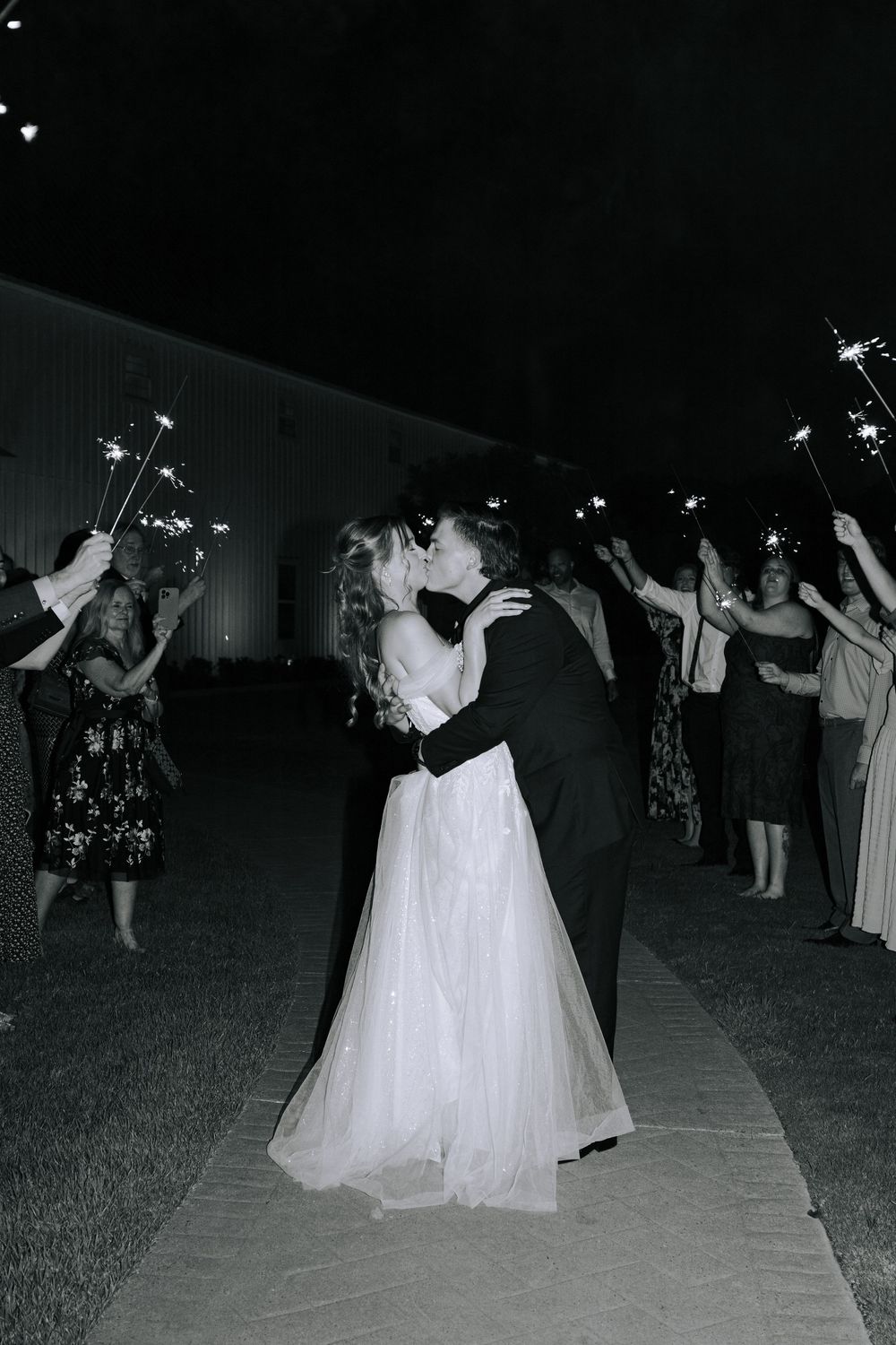Wedding guests hold sparklers during a nighttime celebration with a couple in white and black dancing.