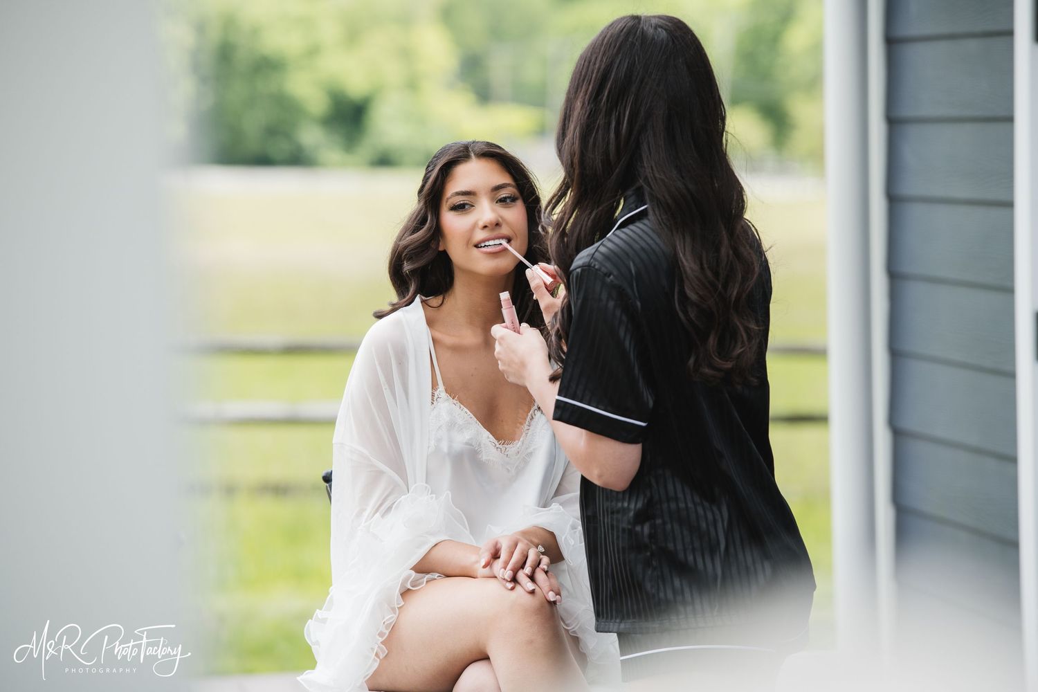 Bride seated outdoors in white robe receiving final makeup touches from her makeup artist.