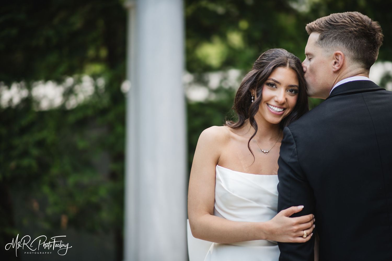 Radiant bride smiles warmly while embracing groom in black tuxedo near white marble columns and greenery.