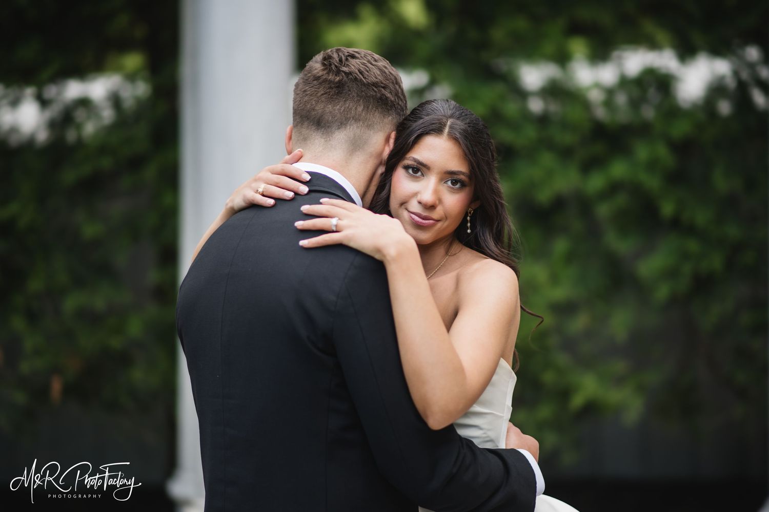 Bride in white strapless wedding gown embraces groom from behind smiling softly in lush garden setting.