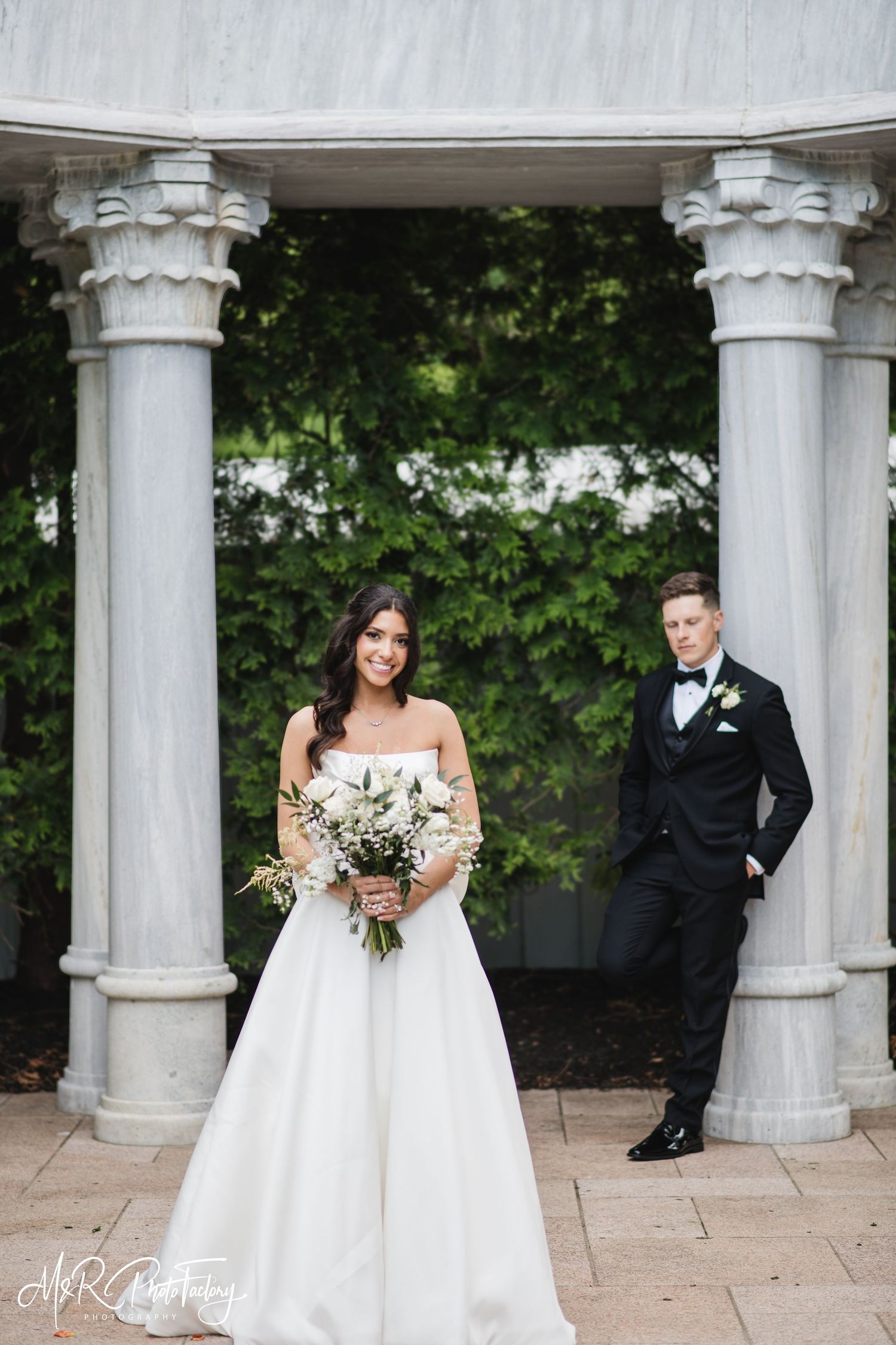 Elegant bride holding white floral bouquet stands before marble gazebo columns as groom poses behind her.
