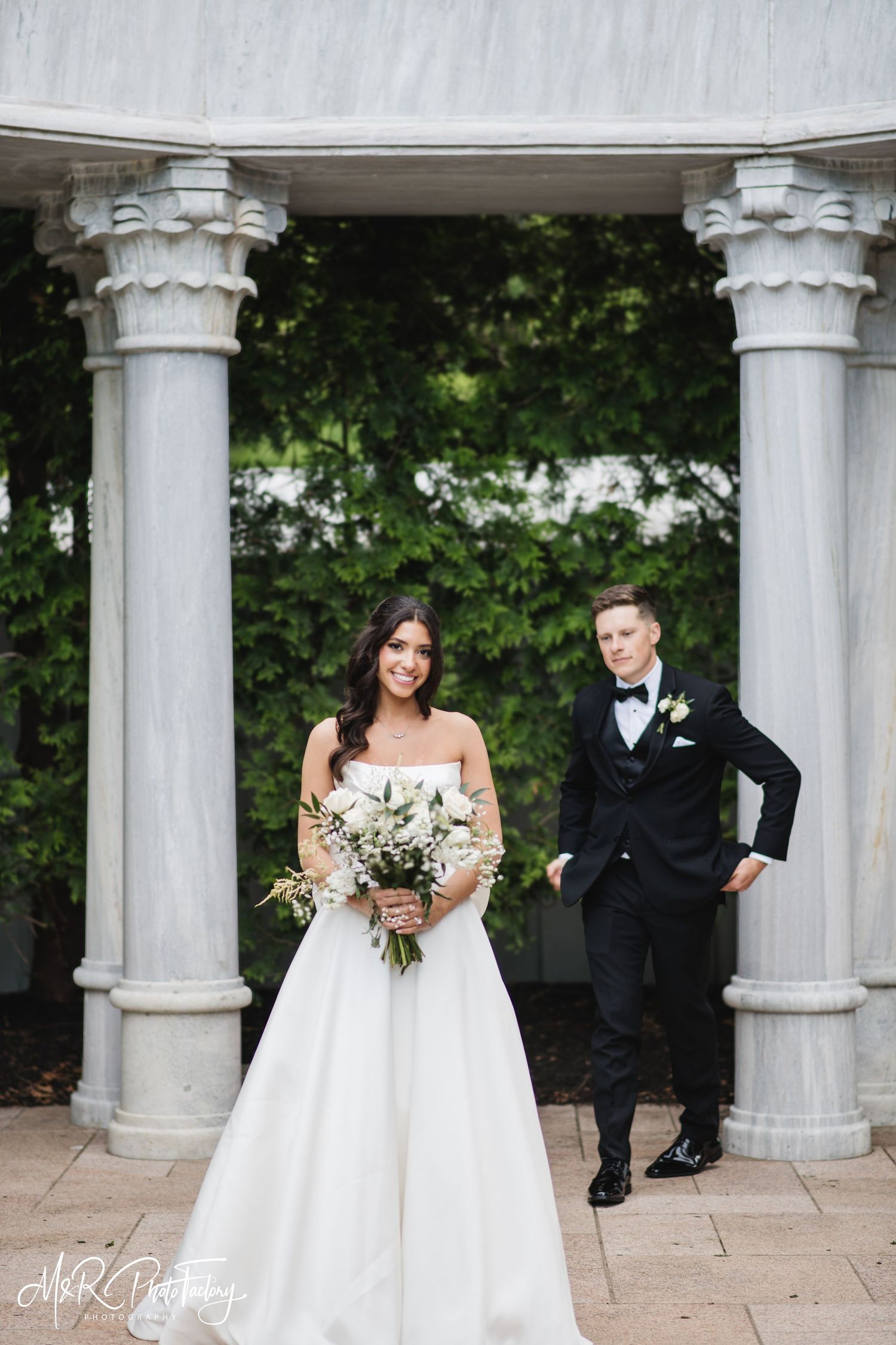 Bride with white bouquet and groom in black tuxedo pose before ornate marble columns with green backdrop.