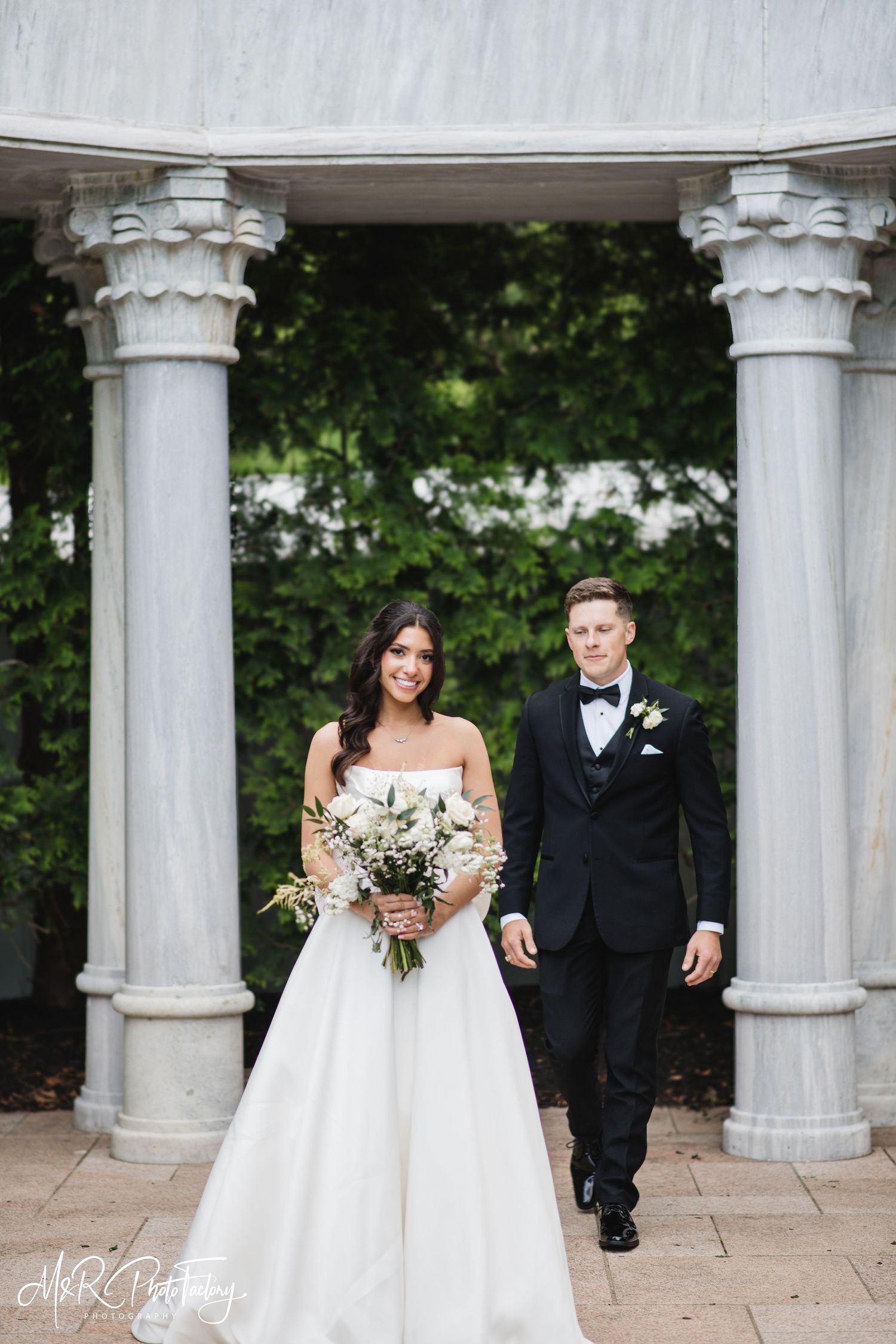 Bride and groom walk hand in hand through marble column gazebo surrounded by lush green foliage.