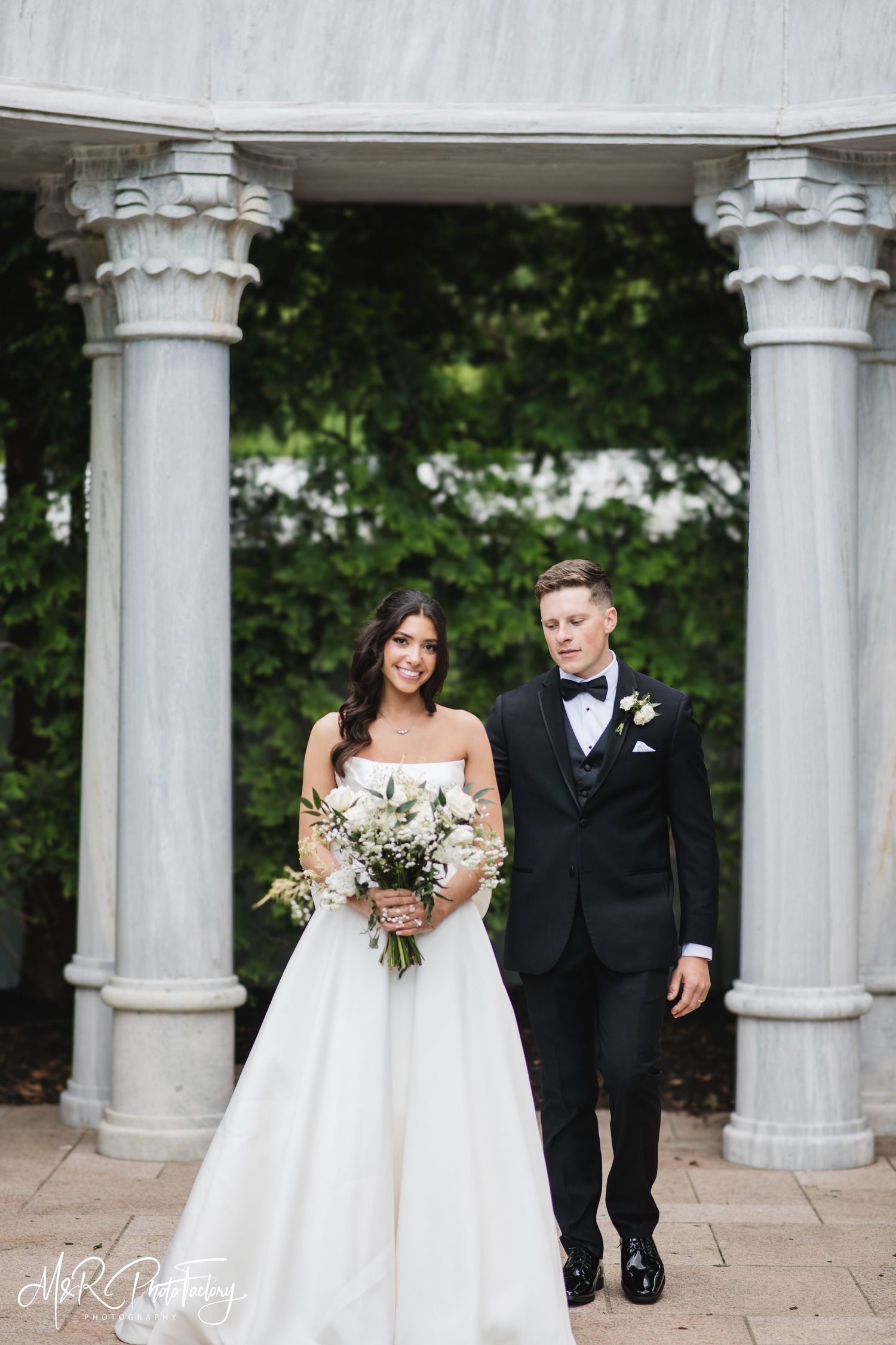 Smiling bride holding white floral bouquet stands with groom in black tuxedo before elegant marble columns.