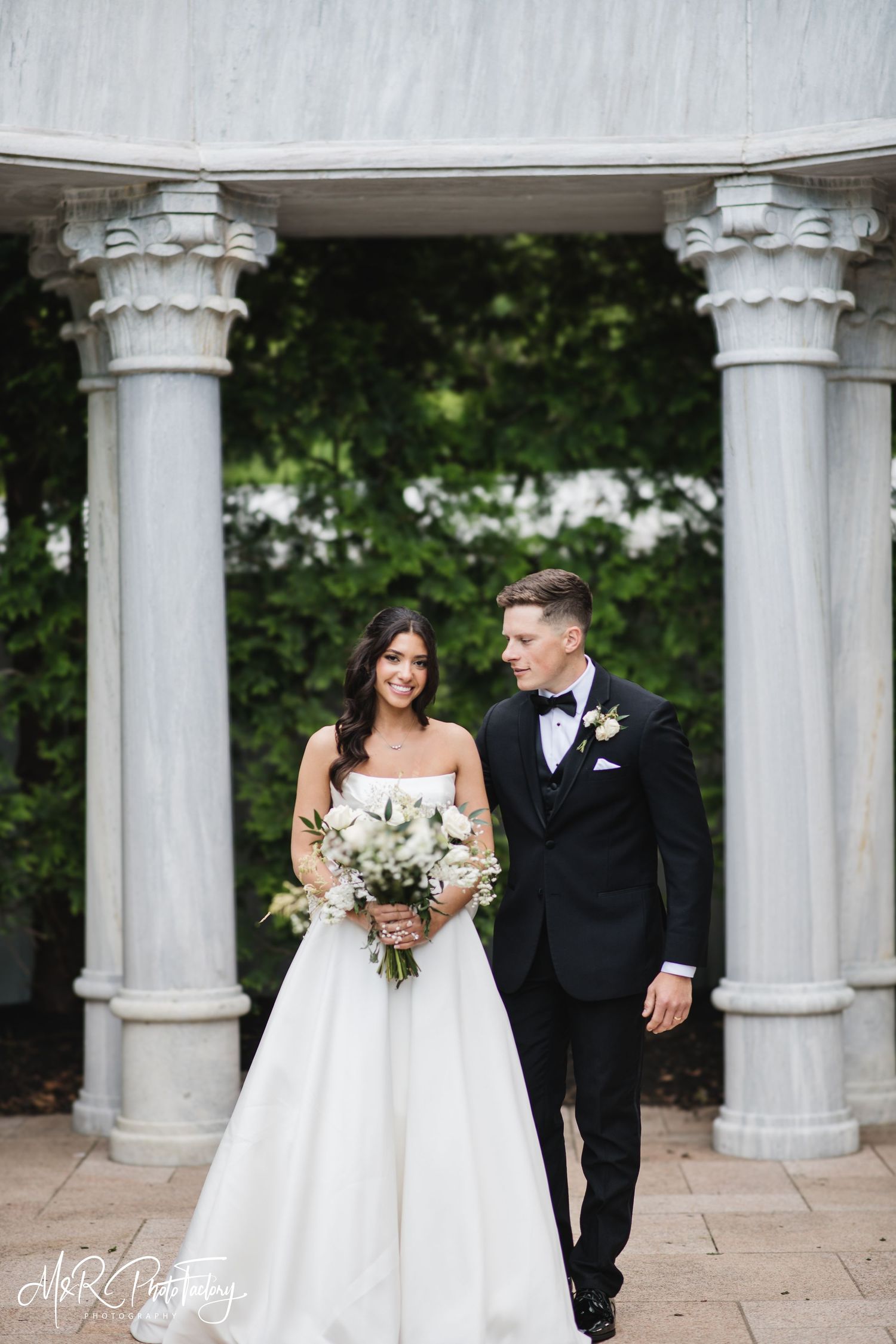 Bride and groom stand before marble columns, she holds white bouquet in elegant strapless ball gown.
