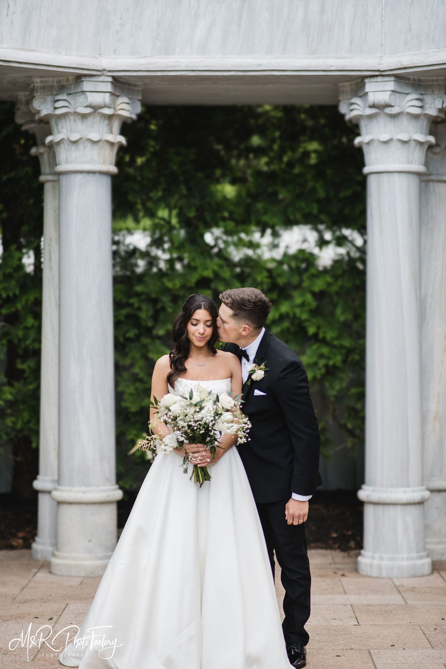 Groom tenderly kisses bride on cheek as she holds white floral bouquet before classic stone columns.