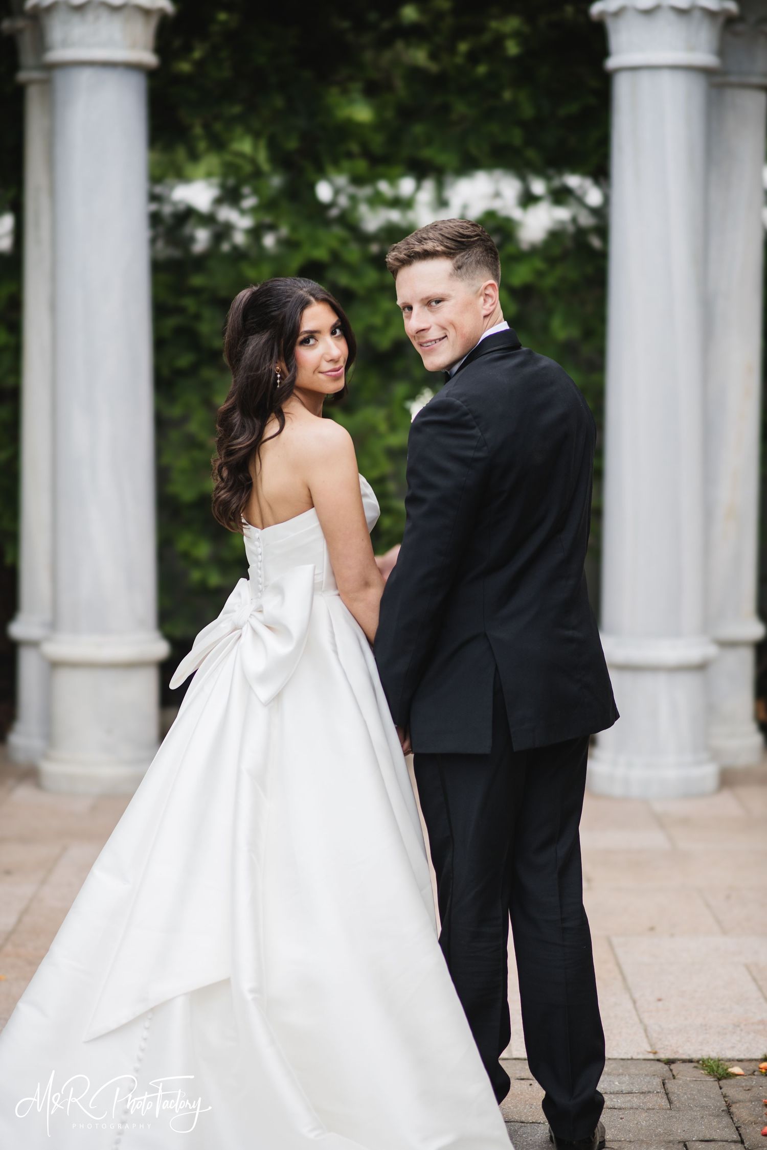 Bride and groom hold hands walking away, she glances back smiling in white bow-back ball gown.