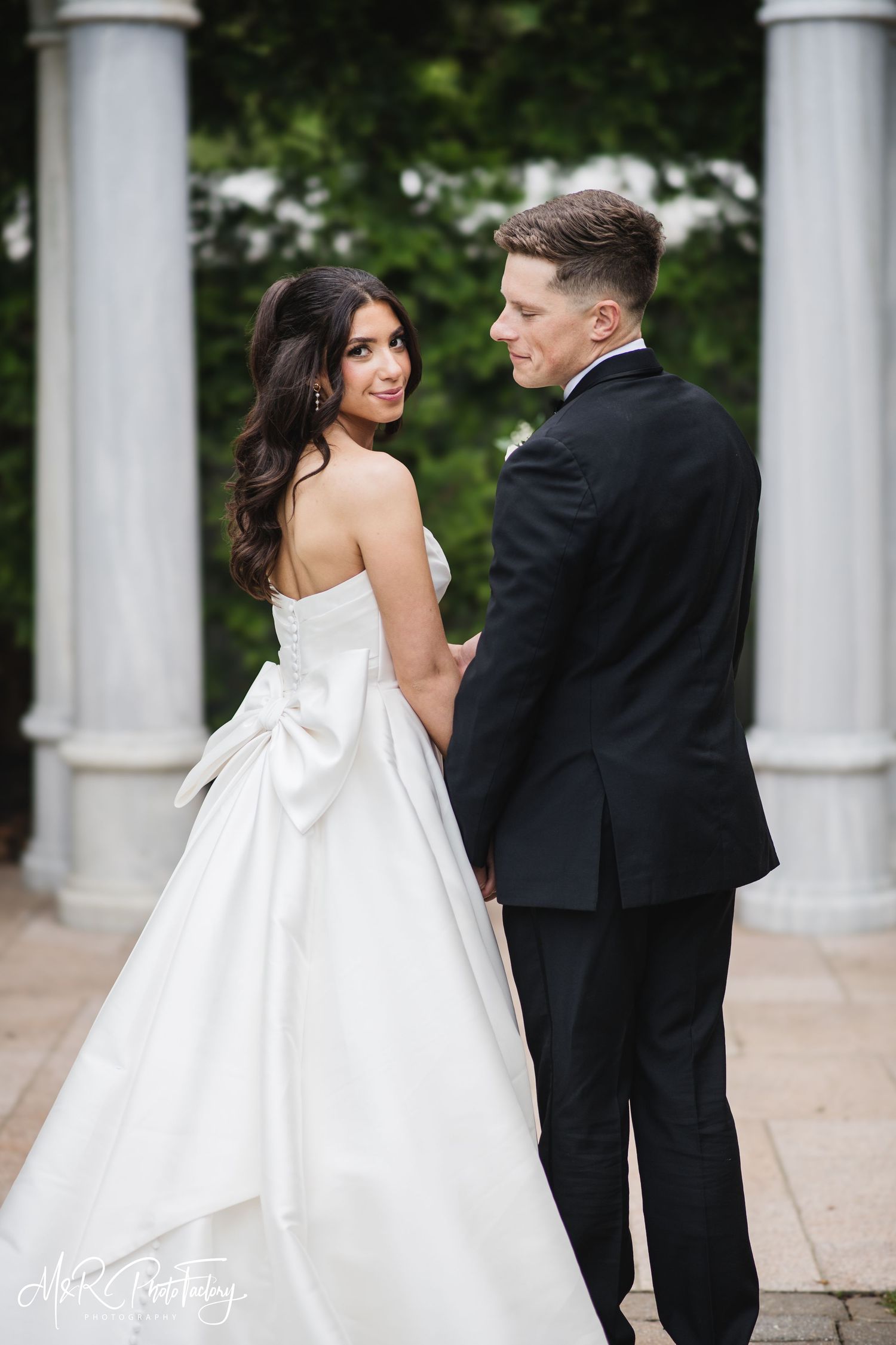 Bride looks over shoulder smiling as groom gazes at her, holding hands near elegant marble columns.