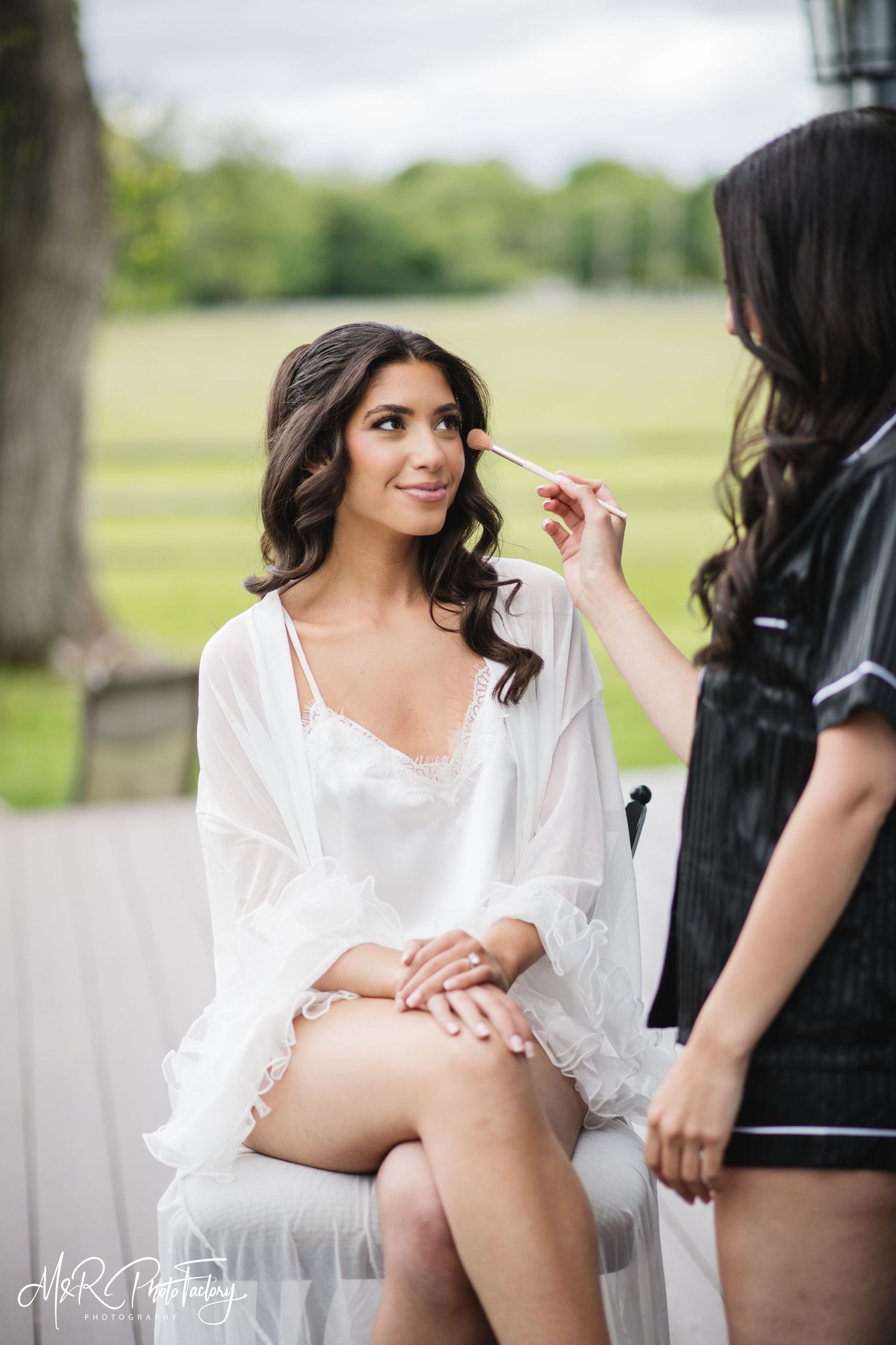 Bride in white lace robe sitting calmly as makeup artist applies blush before her wedding.