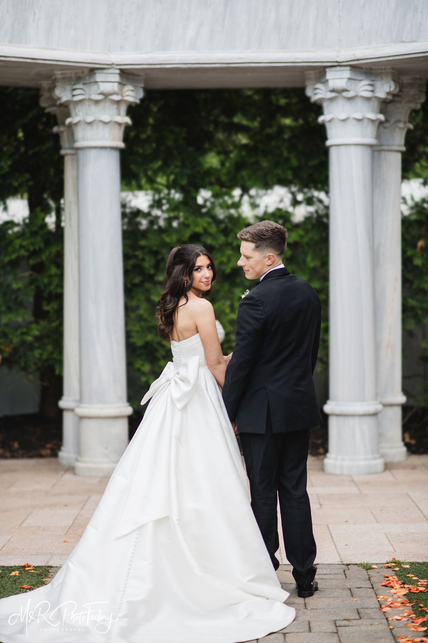 Bride and groom stand facing each other beneath a classical marble columned pavilion on their wedding day.