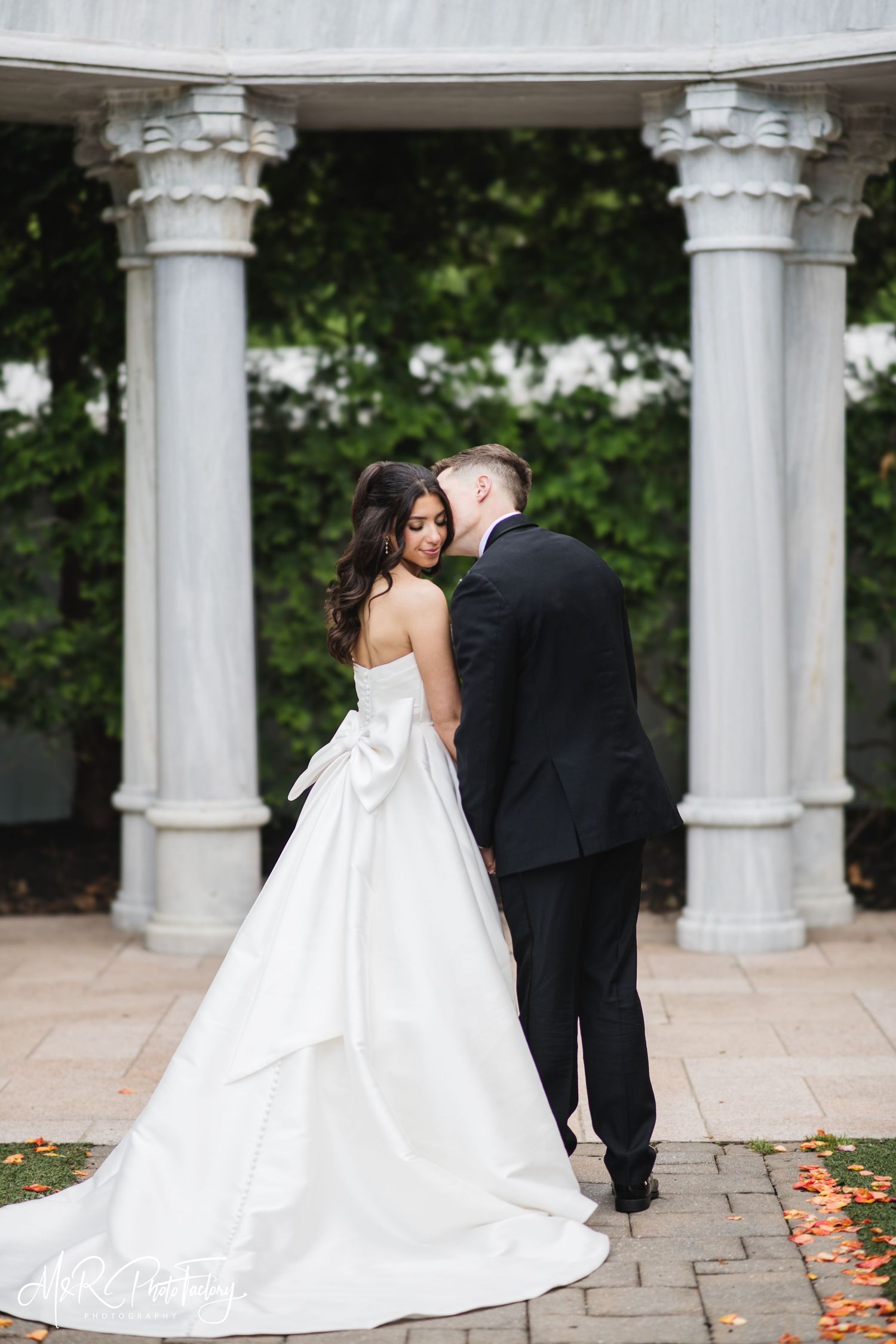 Bride in stunning bow-back white gown and groom share a romantic moment beneath stone columns.