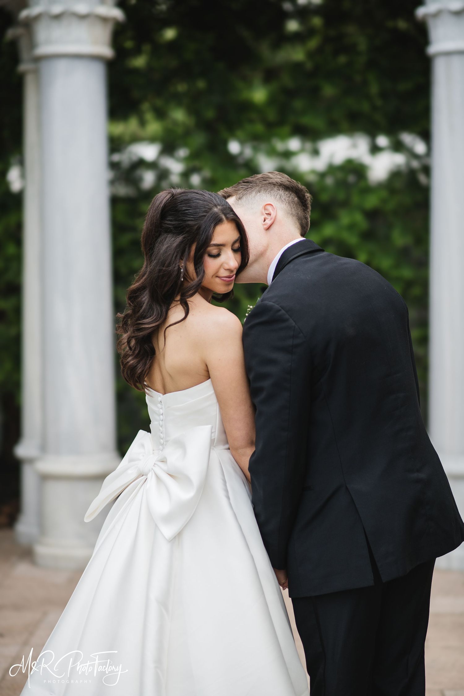 Groom gently nuzzles bride as she looks down, her elegant bow-back wedding gown trailing gracefully.