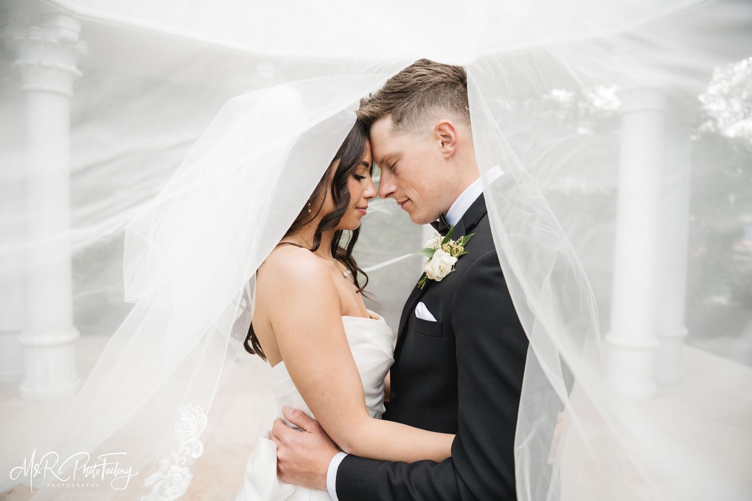 Romantic bride and groom nearly touch foreheads while draped beneath a flowing white wedding veil.