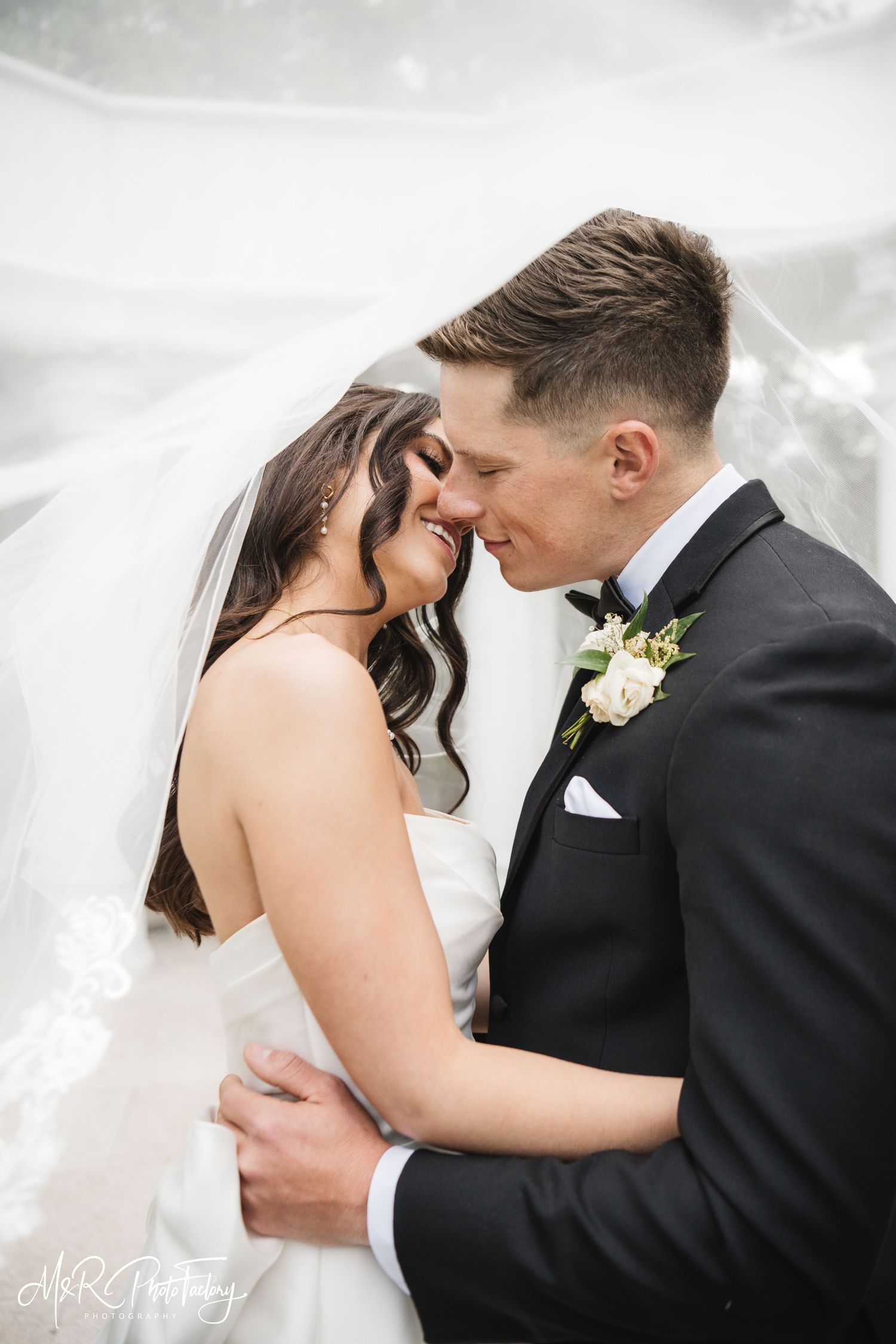 Bride and groom smile intimately under flowing veil, groom in black tuxedo with white boutonniere.