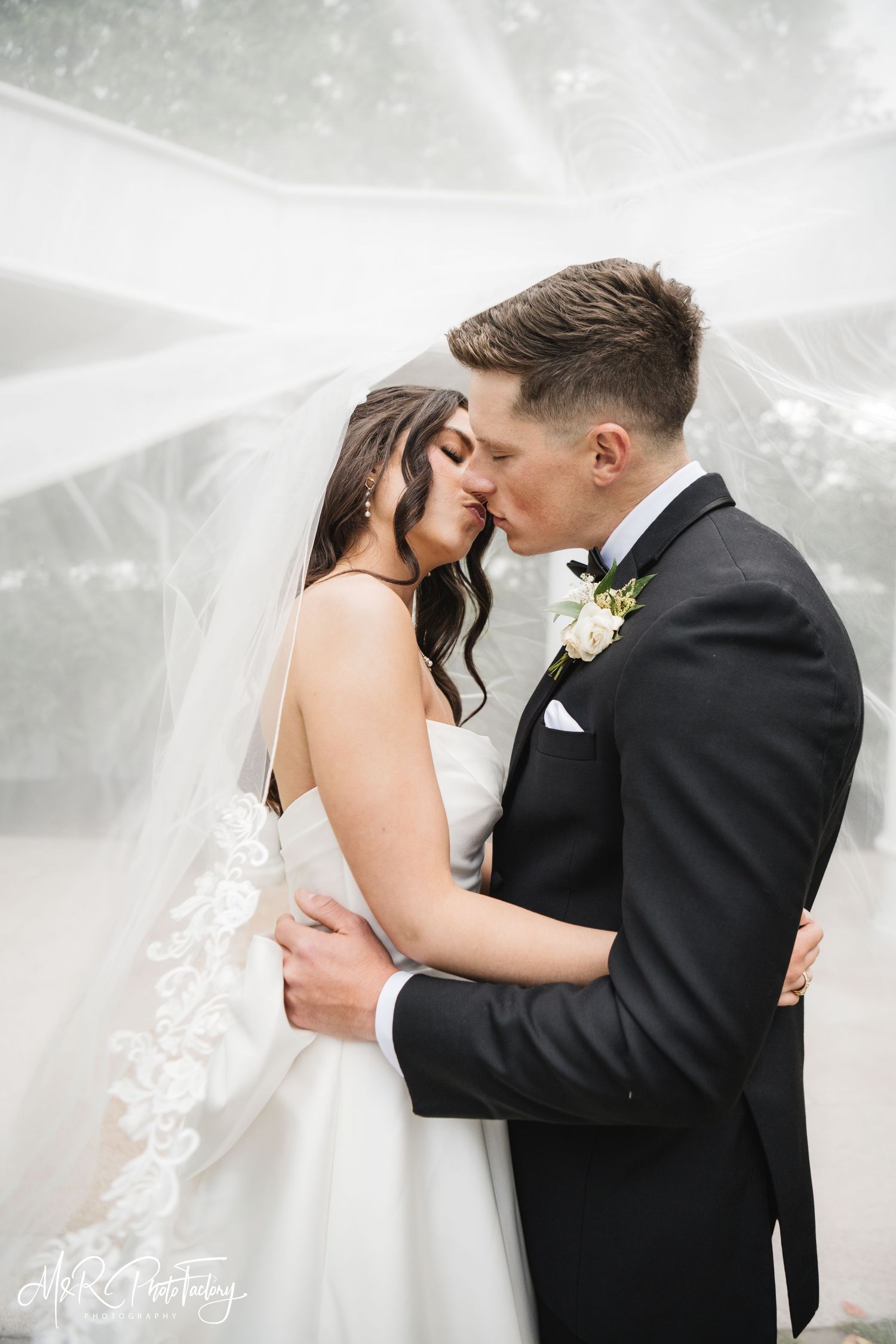 Bride and groom share a tender forehead kiss under a flowing lace veil outdoors.