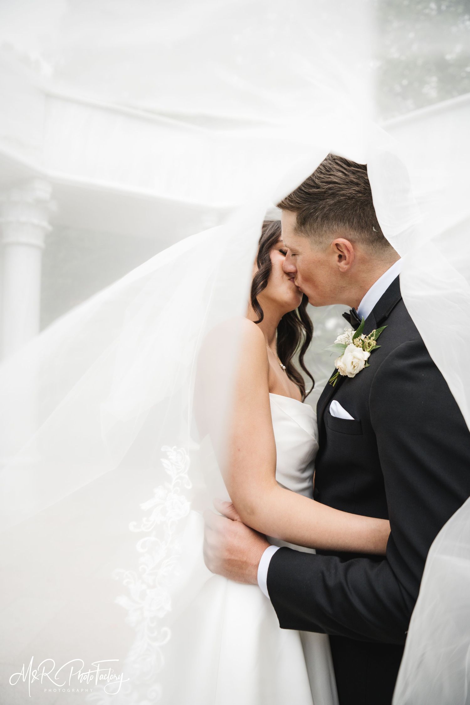 Bride and groom embrace and kiss under a dramatic flowing veil near white columns.