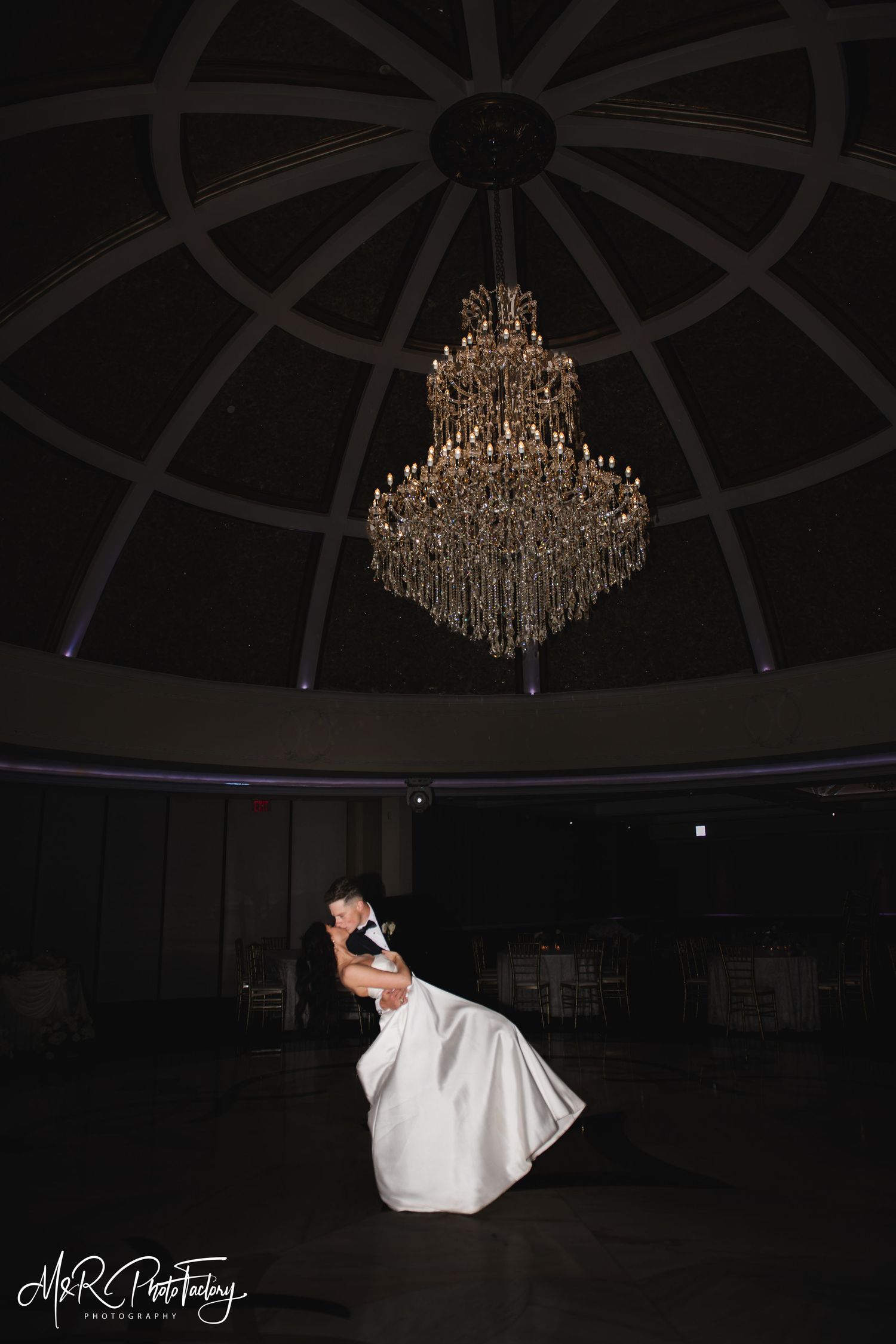 Groom dips bride dramatically under a stunning chandelier in a darkened ballroom.