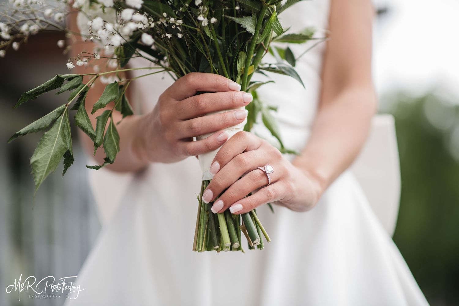 Bride holding a green and white wedding bouquet with manicured nails and a diamond engagement ring.