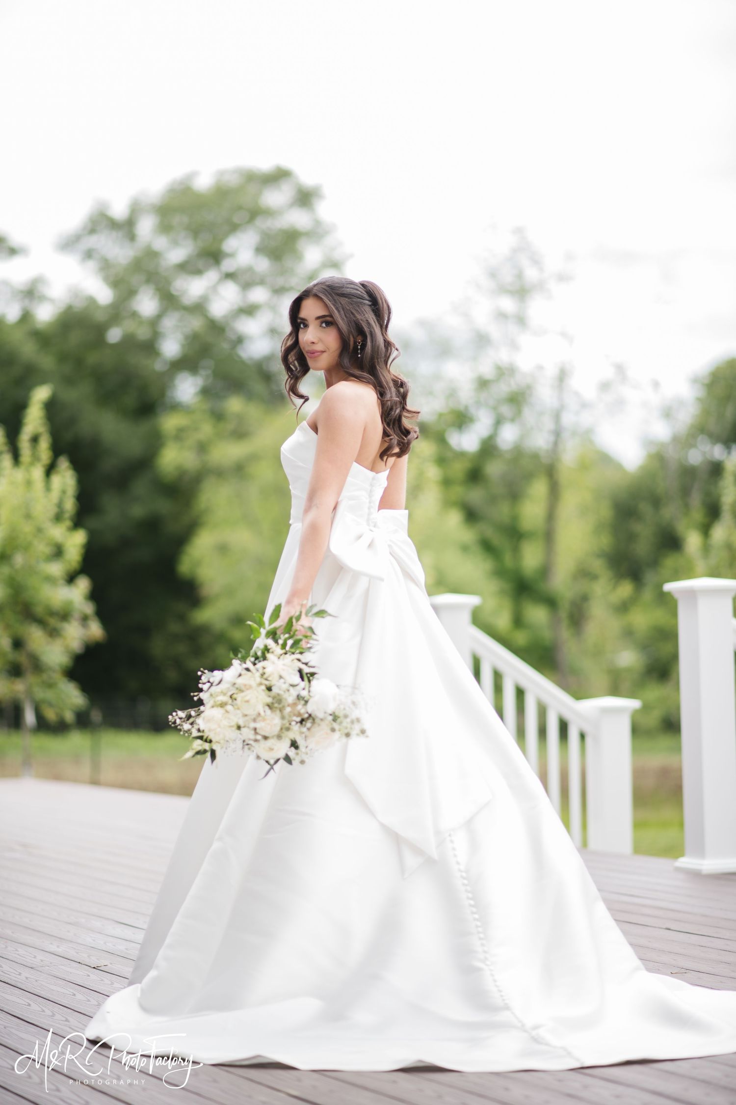 Brunette bride in a strapless white ball gown with bow holding a white bouquet on a deck.
