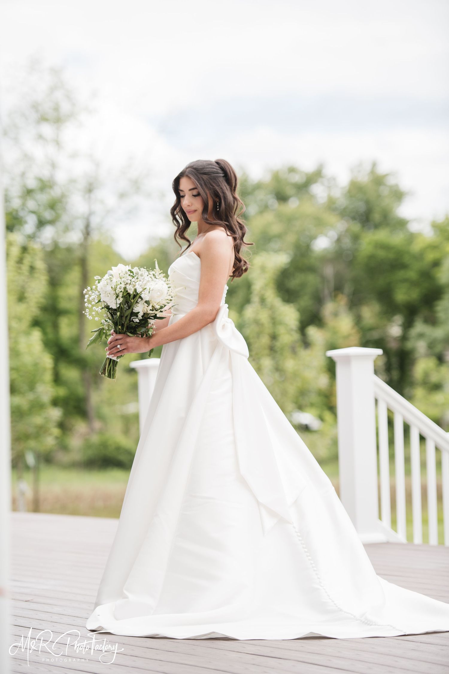 Bride in a flowing white strapless wedding dress holding a white bouquet on an outdoor deck.