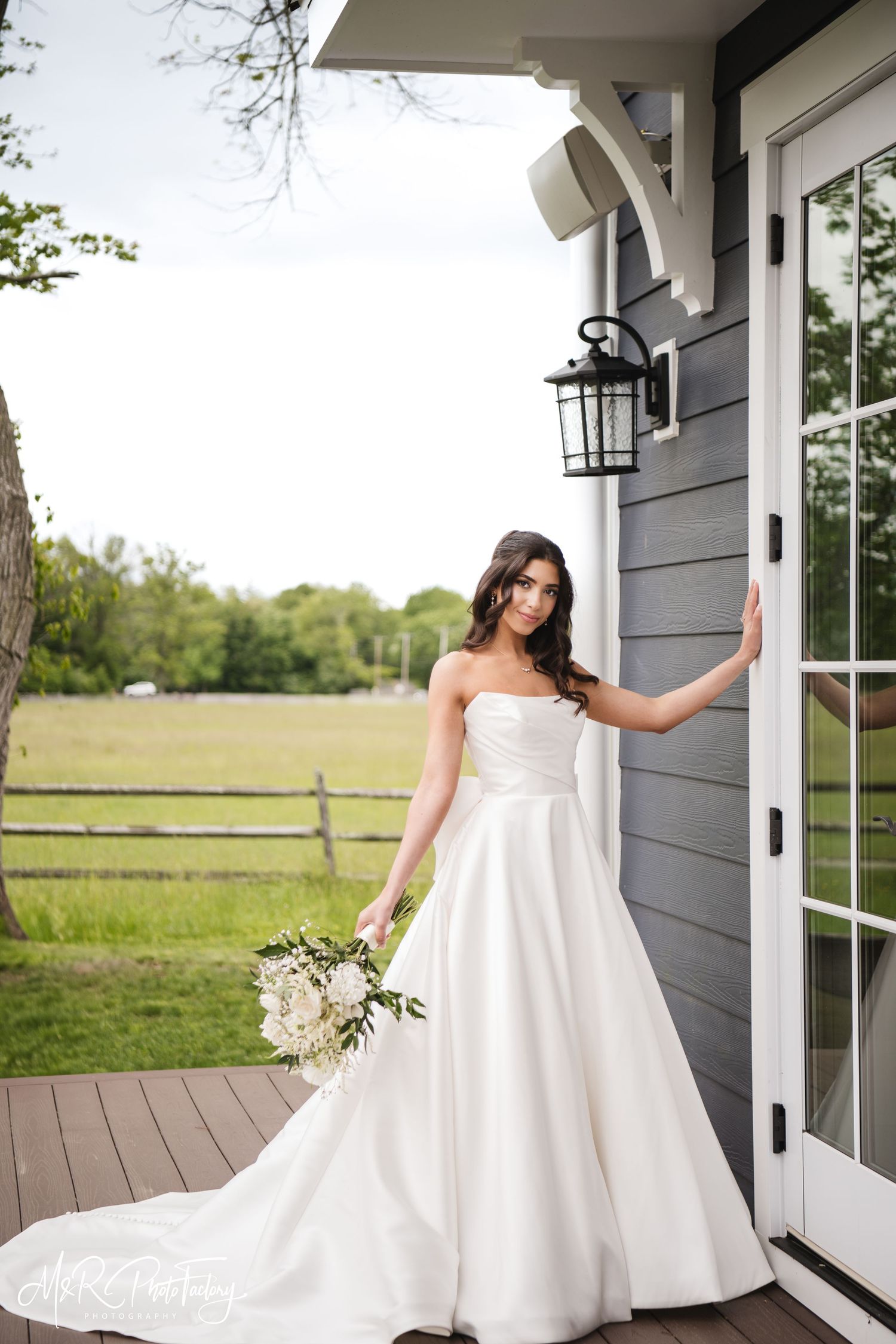 Elegant bride in a strapless white gown posing by a gray farmhouse door with white bouquet.