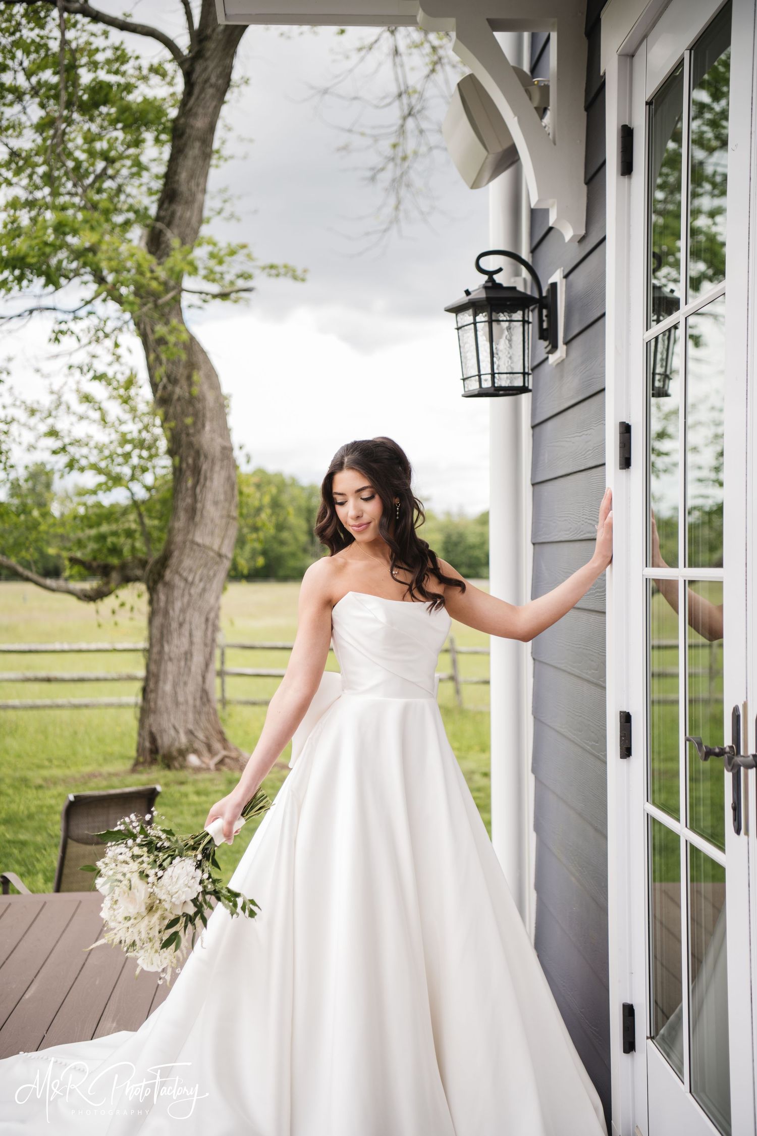 Bride in a strapless white ball gown looking down while holding bouquet near a farmhouse door.