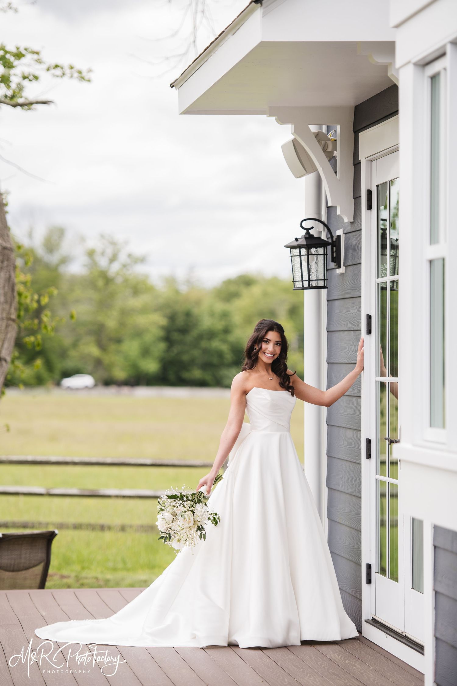 Beautiful bride in a strapless white wedding gown posing on a porch of a gray farmhouse.
