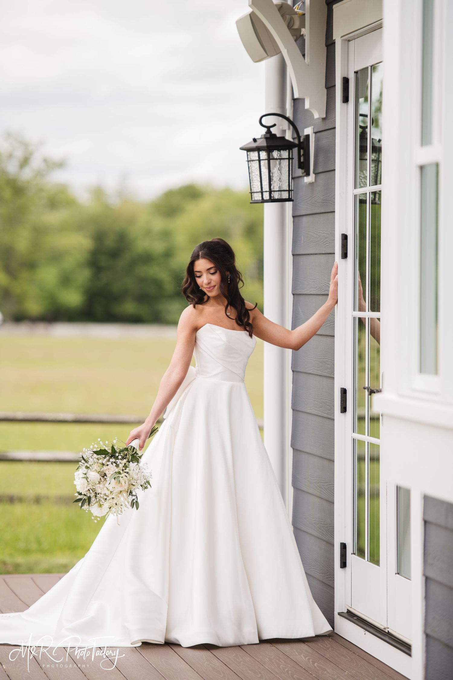 Stunning bride in a white strapless ball gown holding a bouquet beside a gray farmhouse door.