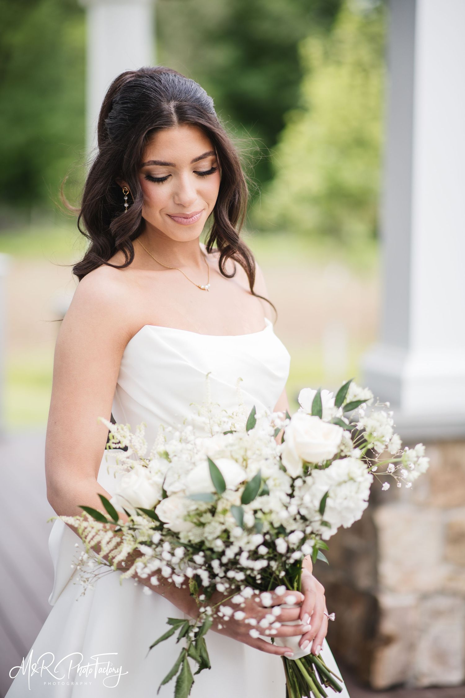 Bride in strapless white gown holding elegant white rose and greenery bouquet with baby's breath outdoors.