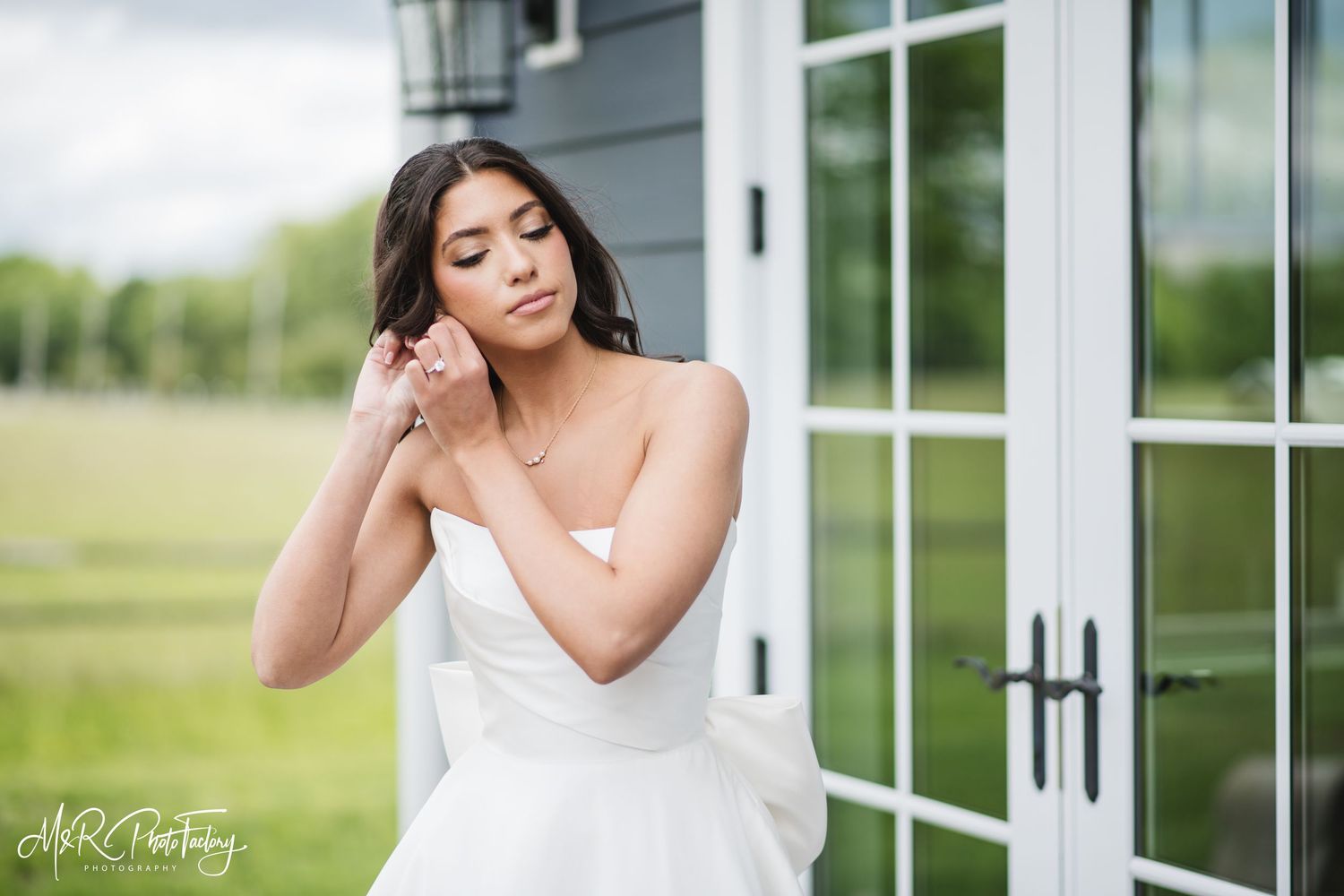 Beautiful bride in strapless white wedding dress adjusting earring beside glass French doors outdoors.