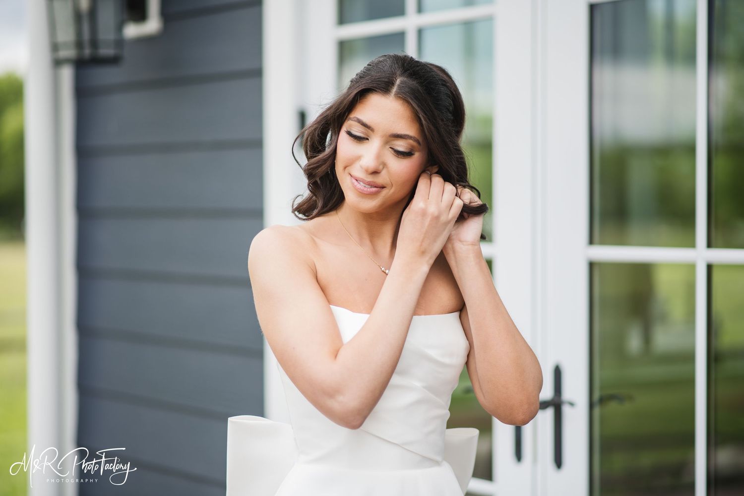 Elegant bride in strapless white gown touching her earring while seated outside near French doors.