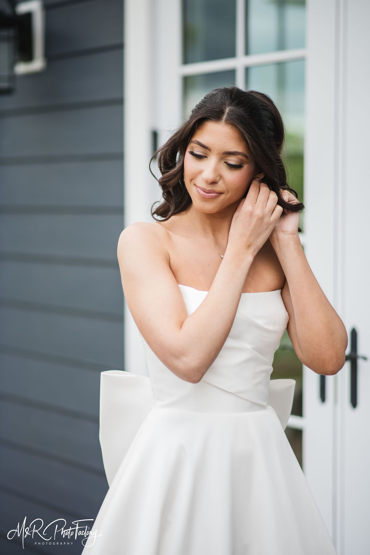 Stunning bride in strapless white ballgown with large bow adjusting earring outside on wedding day.