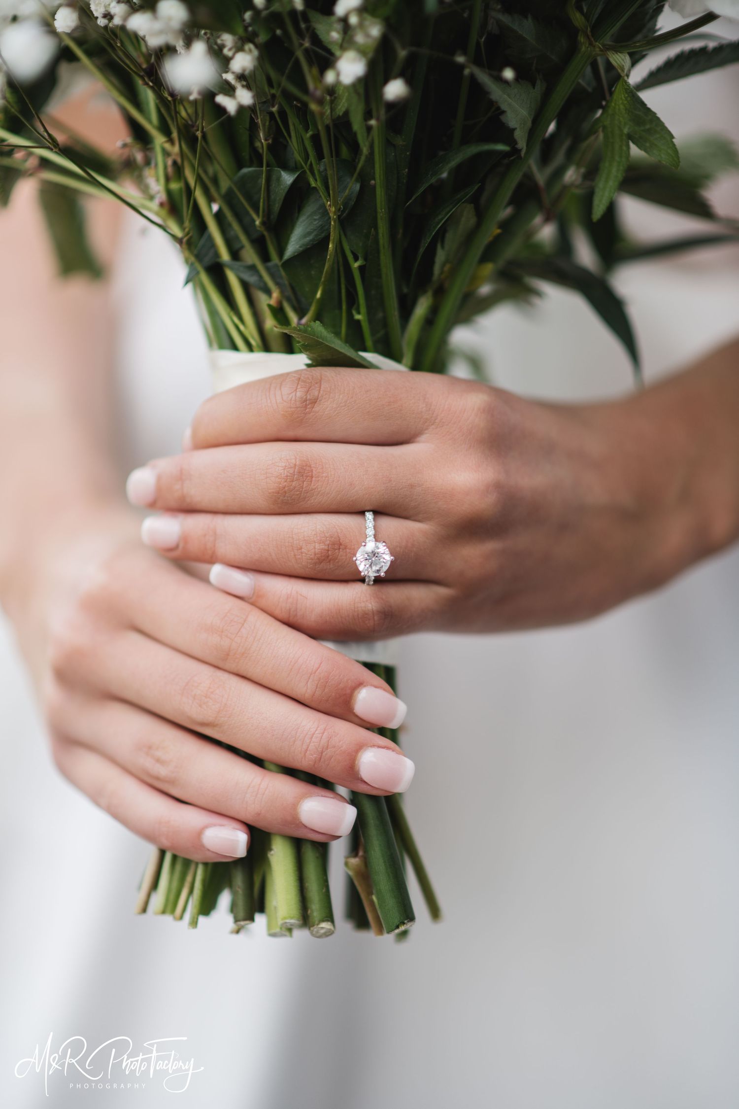 Close-up of bride's hands holding green bouquet stems showcasing solitaire diamond engagement ring.