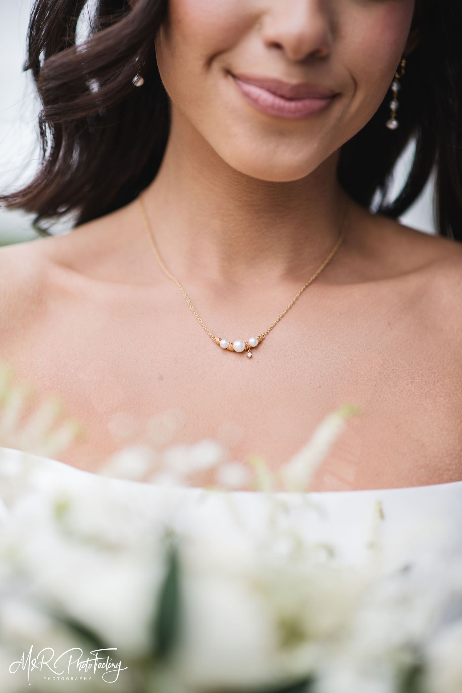 Close-up of bride wearing delicate gold pearl necklace and drop earrings holding white floral bouquet.