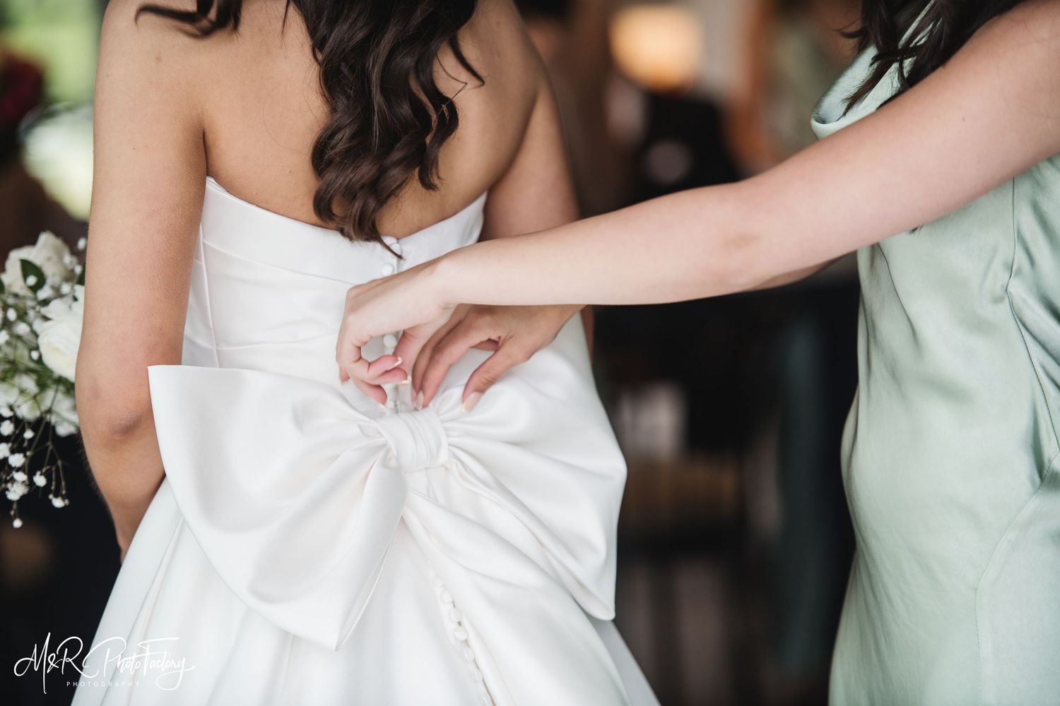 Bridesmaid in sage green dress fastening large bow on back of bride's white strapless wedding gown.