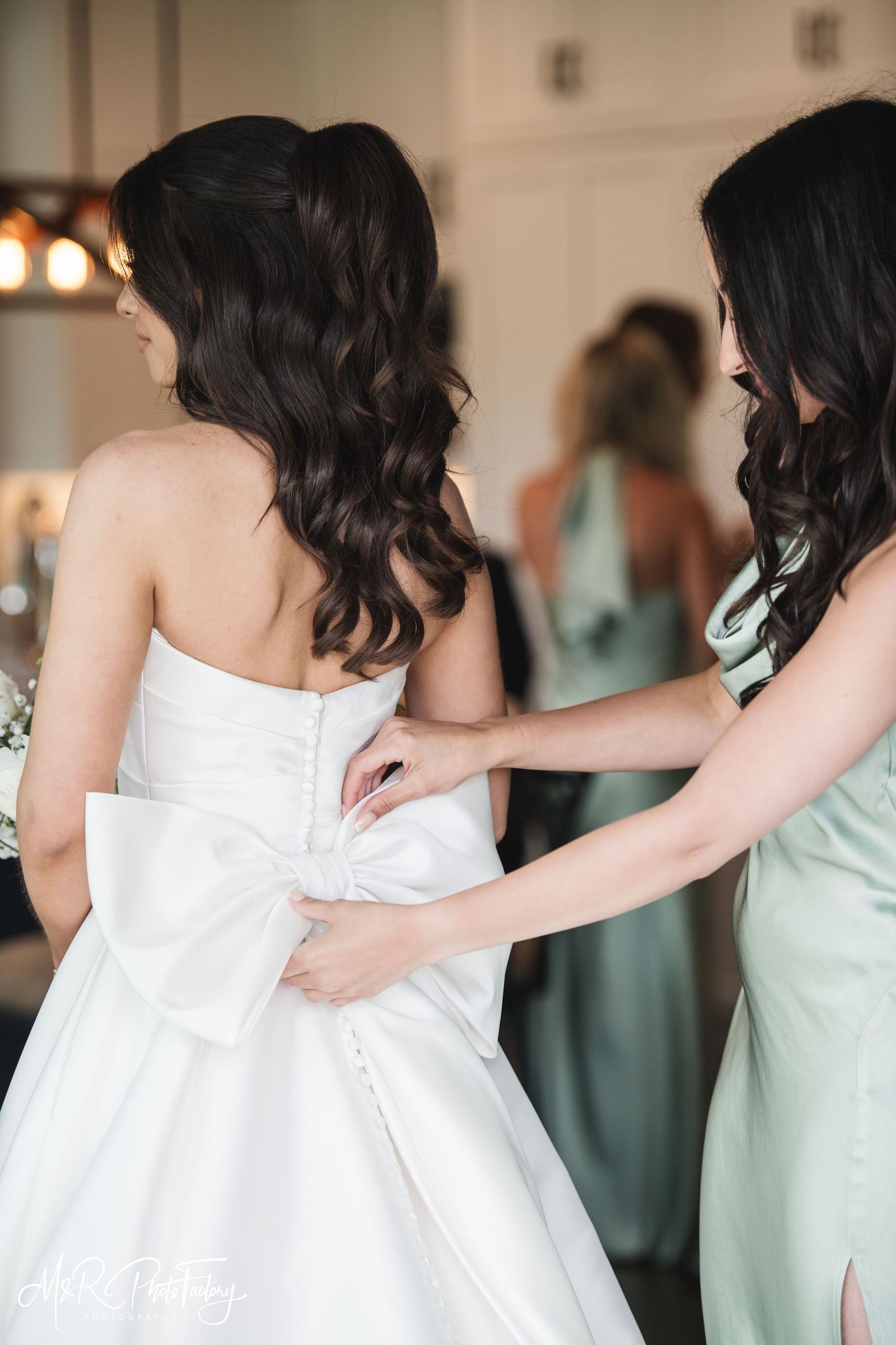 Bridesmaid helping button the back of bride's white strapless ballgown with oversized bow detail indoors.