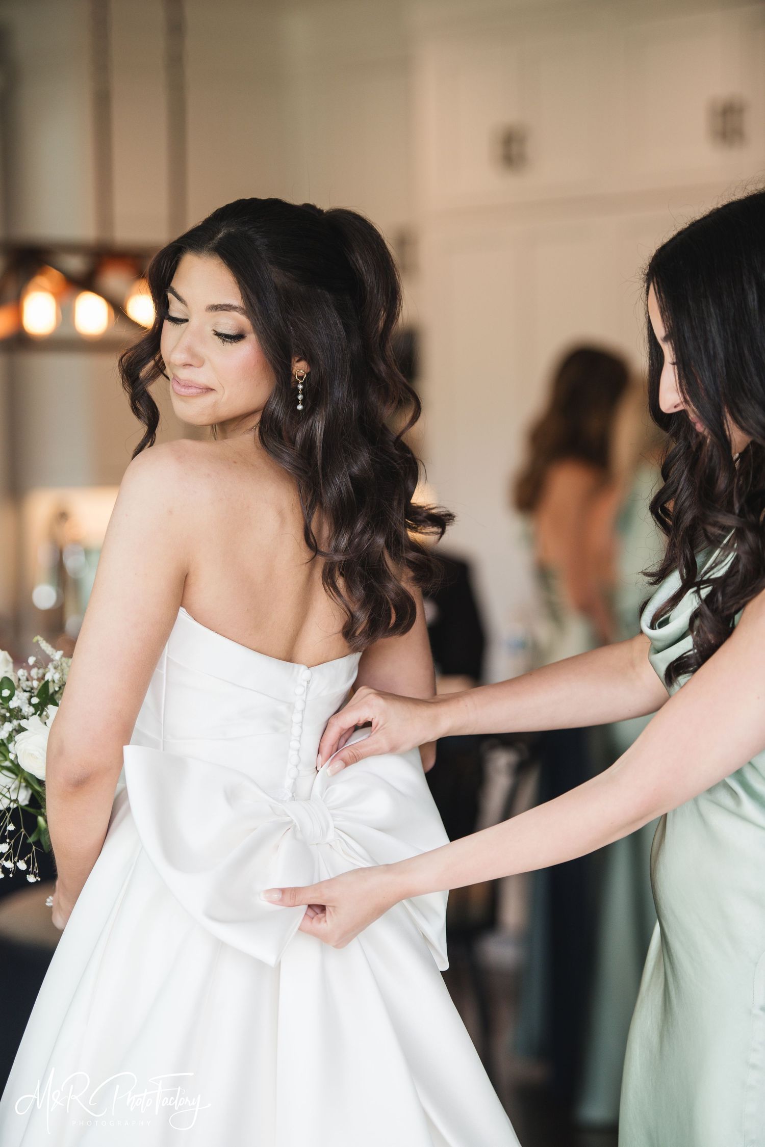 Bridesmaid in mint dress buttoning bride's white strapless gown with large bow in bright bridal suite.
