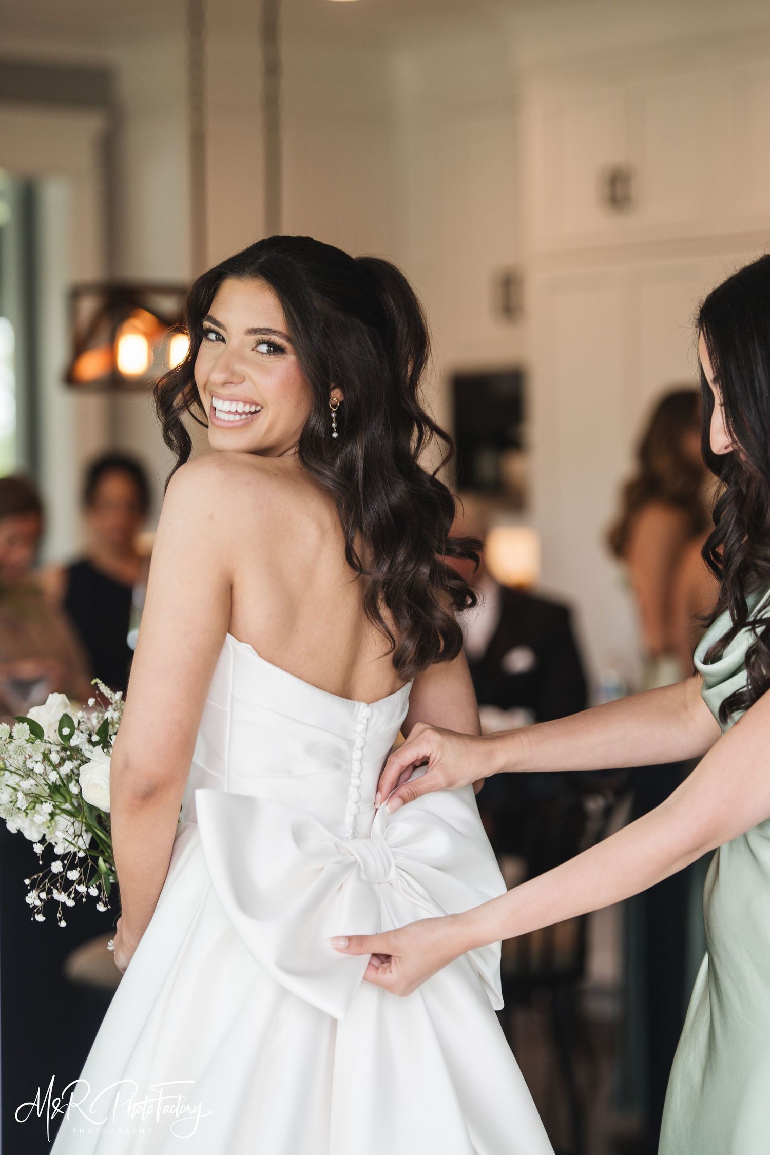 Bride smiling over shoulder as bridesmaid fastens large bow on back of strapless white wedding gown.