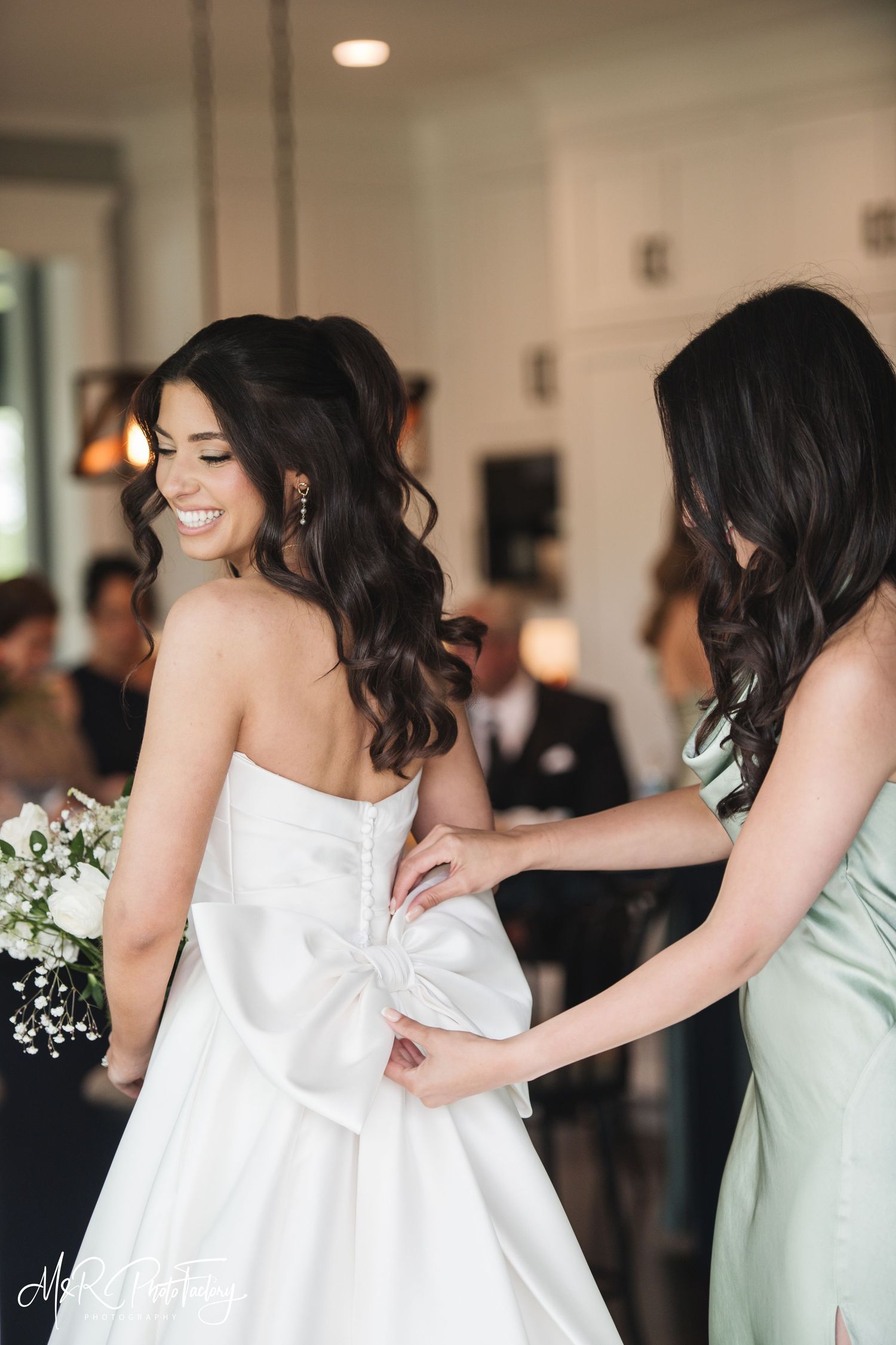 Bridesmaid in sage green dress buttoning the back of bride's elegant strapless ball gown with bow detail.