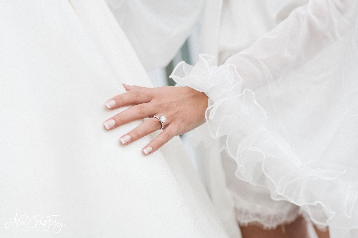 Close-up of bride's hand with delicate engagement ring resting on white ruffled wedding dress fabric.